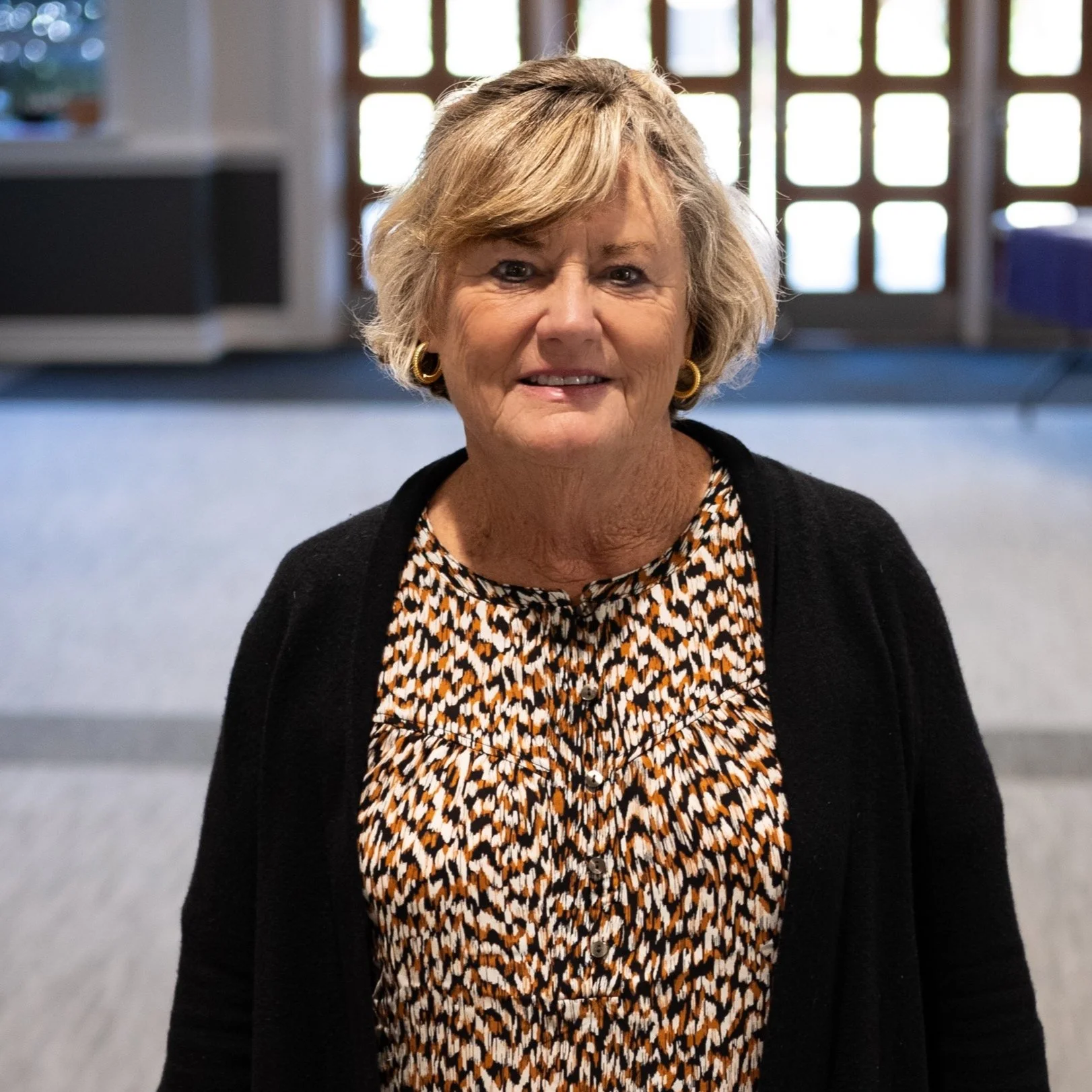 An elderly woman with blonde hair and gold earrings, wearing a black cardigan over a patterned blouse, standing indoors with a window in the background.