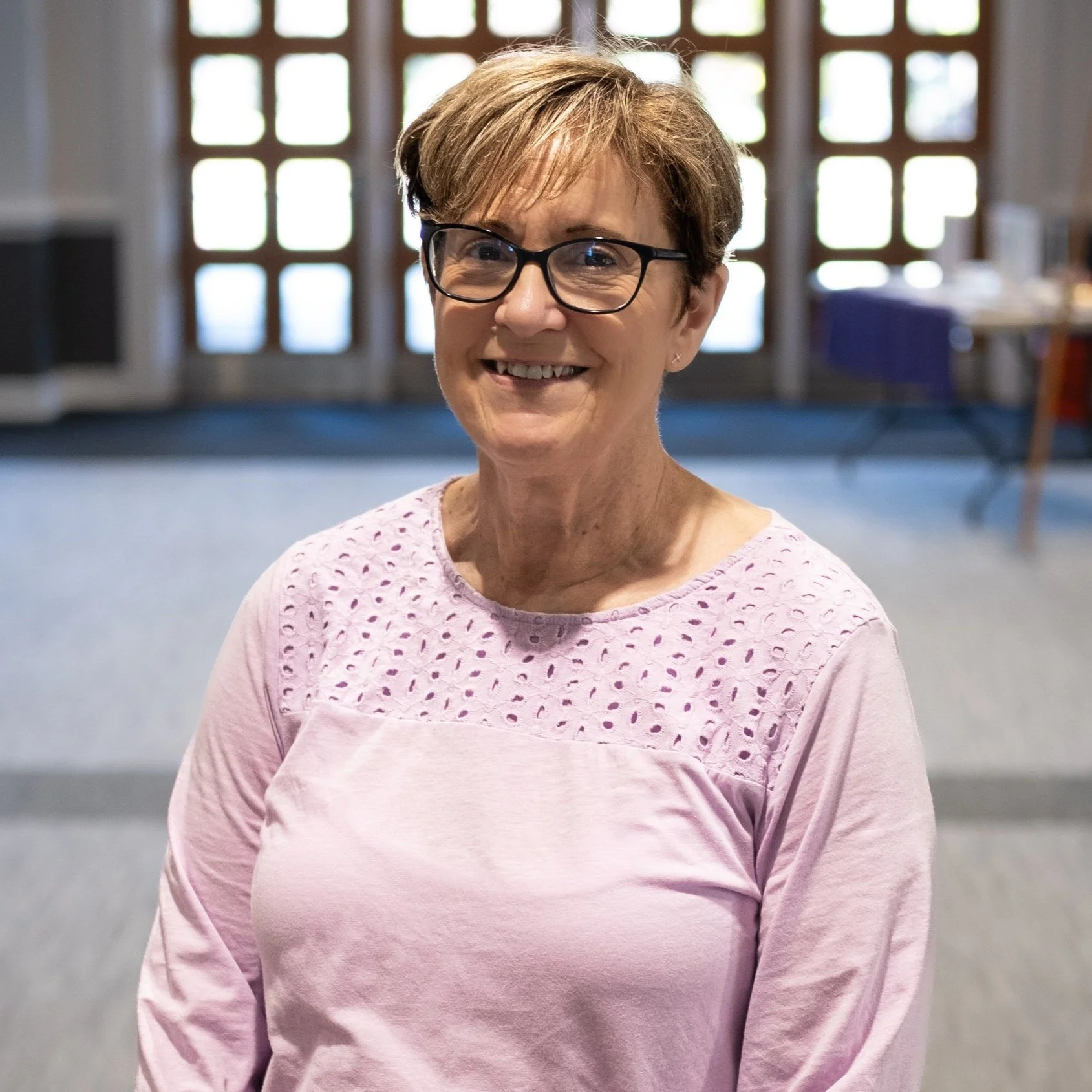 An older woman with short light brown hair, glasses, and a pink shirt with eyelet details on the shoulders, smiling indoors in front of large windows.