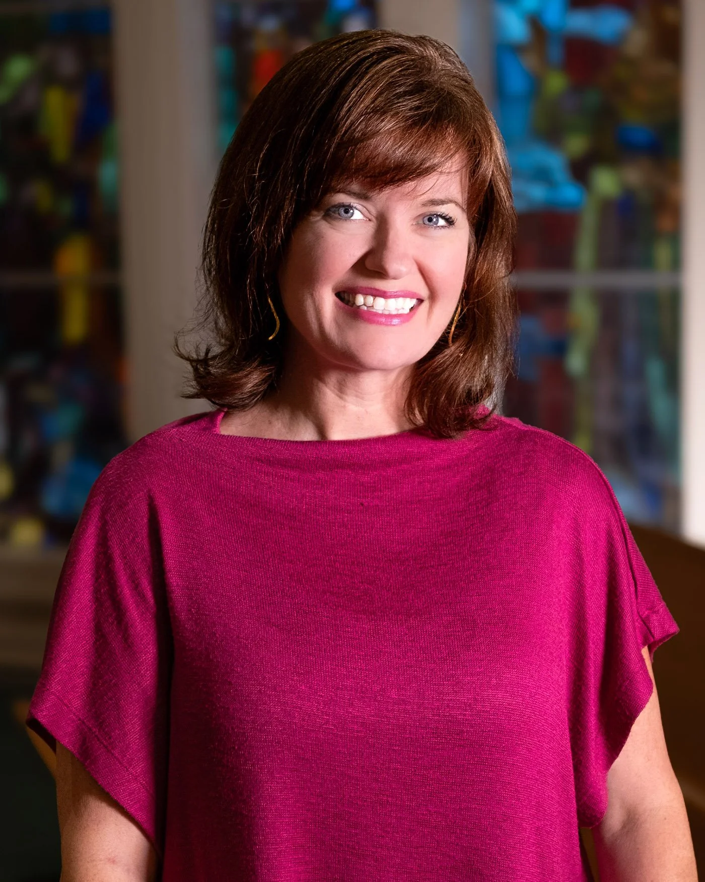A woman with shoulder-length brown hair, smiling, wearing a pink top, standing indoors with colorful artwork or decorations in the background.
