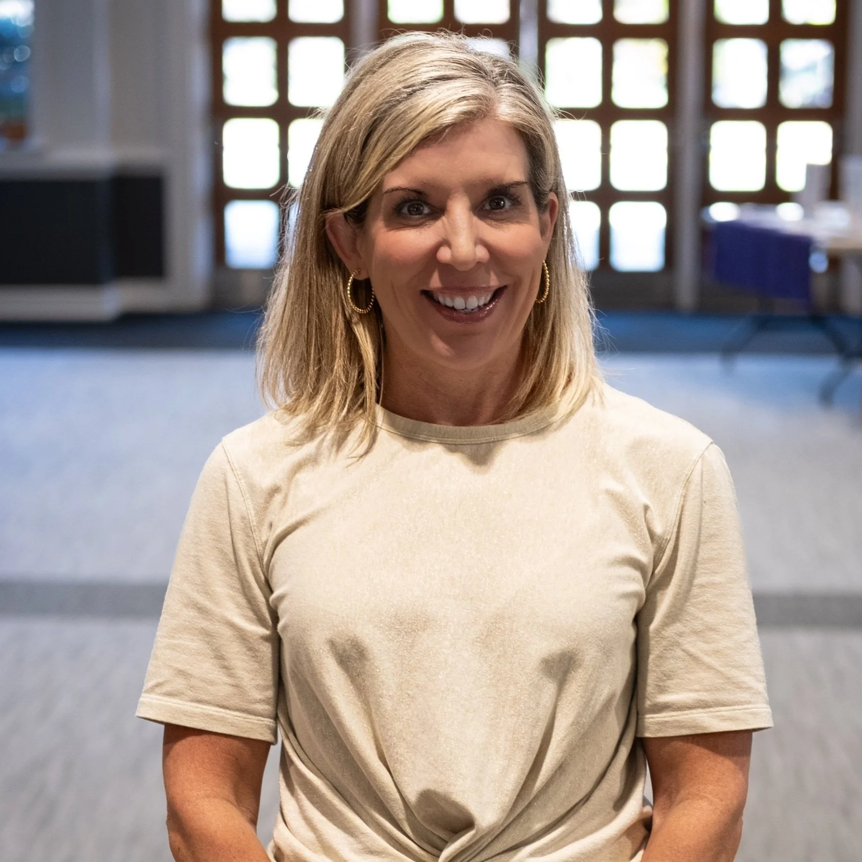 A smiling woman with shoulder-length blonde hair wearing a beige t-shirt and gold hoop earrings, standing inside a building with large windows and a door in the background.
