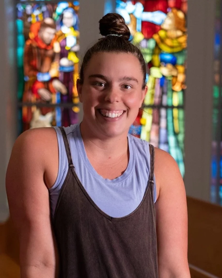 Young woman with a top knot hairstyle, smiling, wearing a beige textured fleece jacket inside a room with large windows.