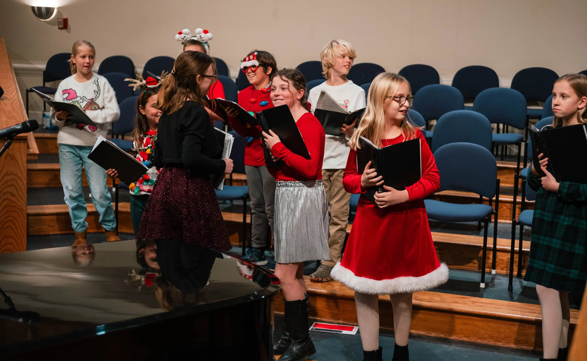 Children in holiday costumes practicing or performing a choir on stage with Christmas decorations.