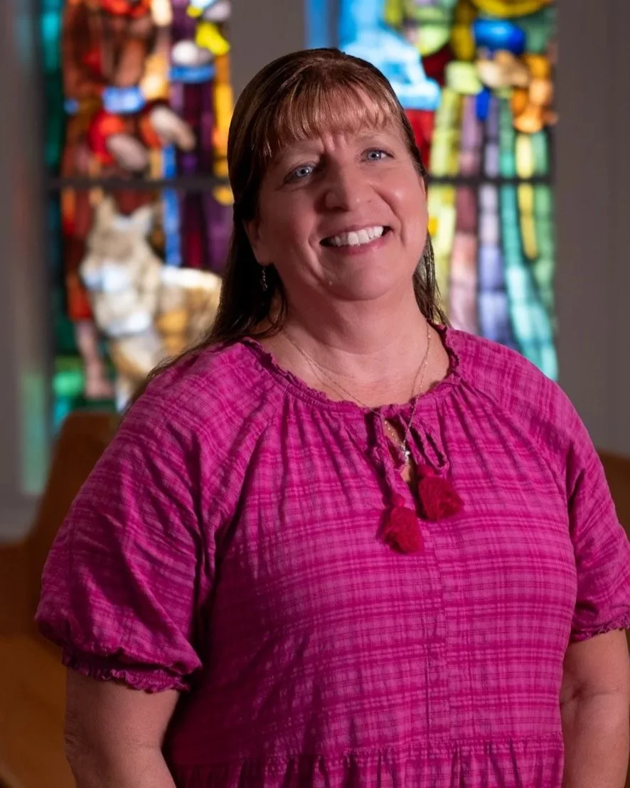 A woman with light brown hair, wearing a black shirt, smiling inside a room with a patterned window in the background.