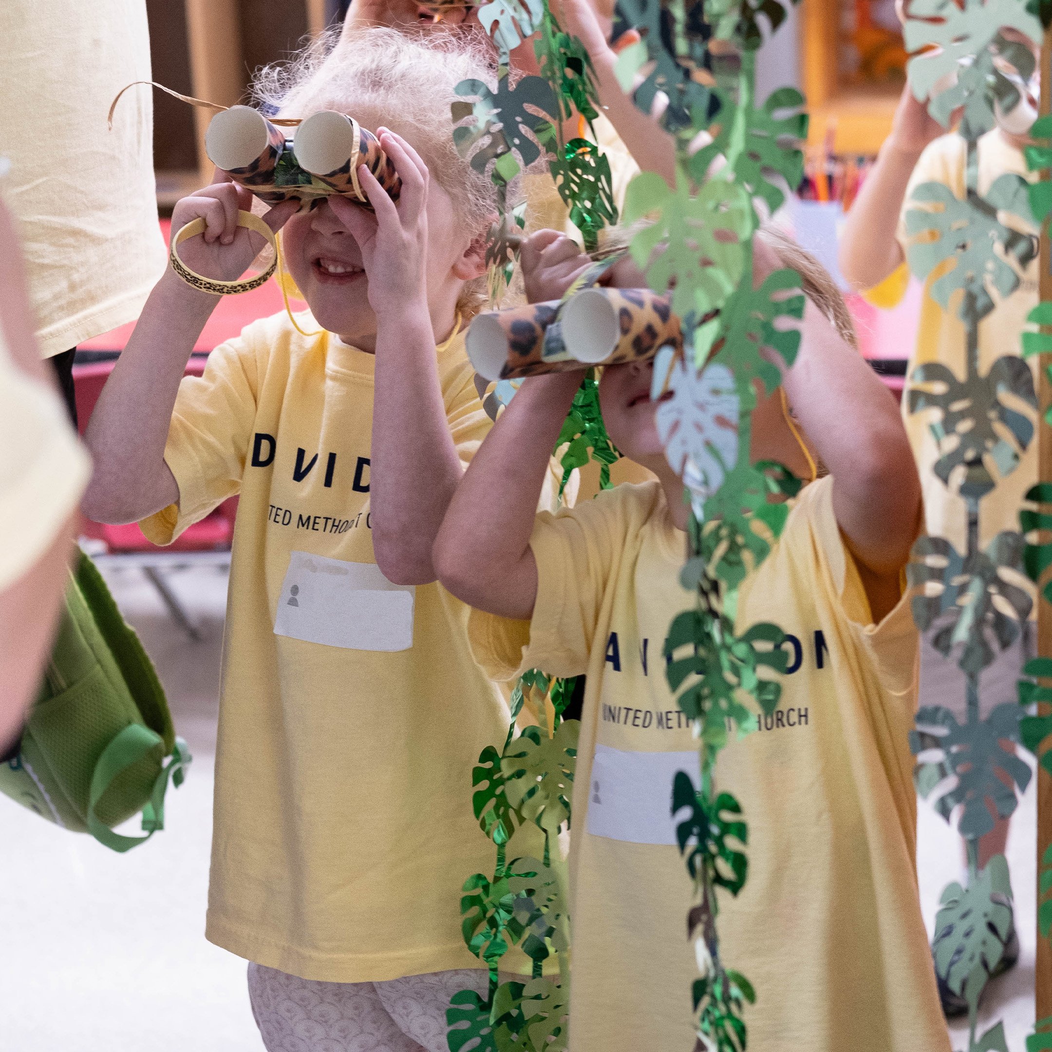 Two children wearing yellow T-shirts with text, peering through cardboard tubes in a creative, jungle-themed setting with hanging paper leaves.