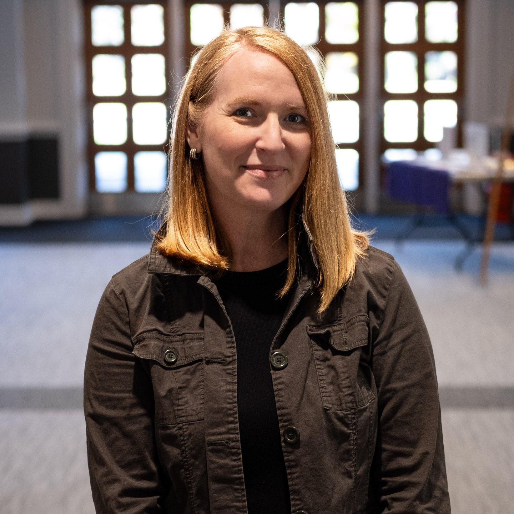 A woman with shoulder-length red hair, wearing a black jacket, smiling indoors in front of large windows.