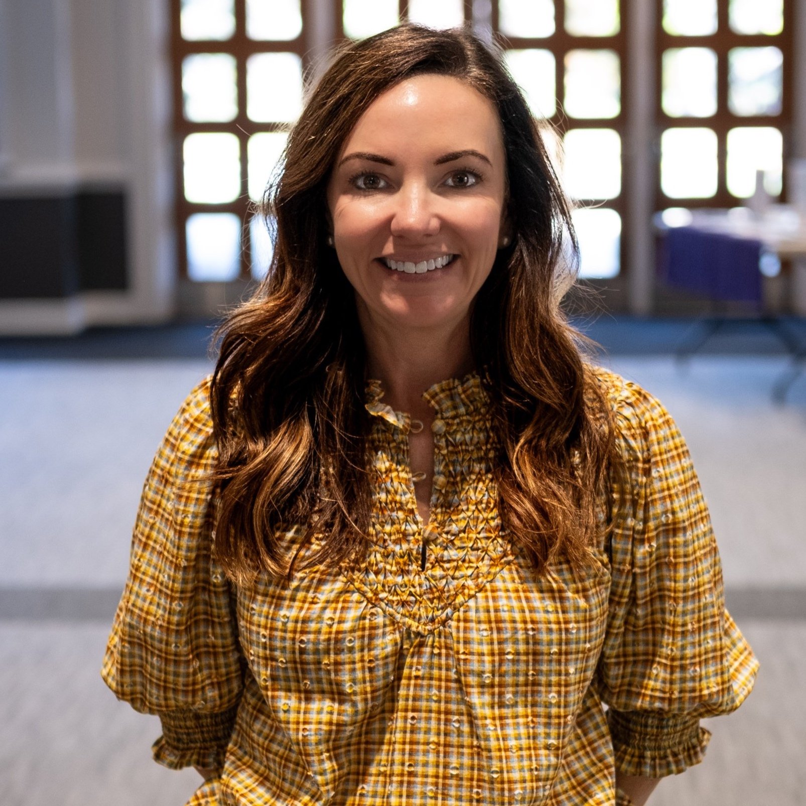 Close-up of a woman with long brown hair, wearing a yellow plaid shirt, smiling, indoors with natural light coming through windows.