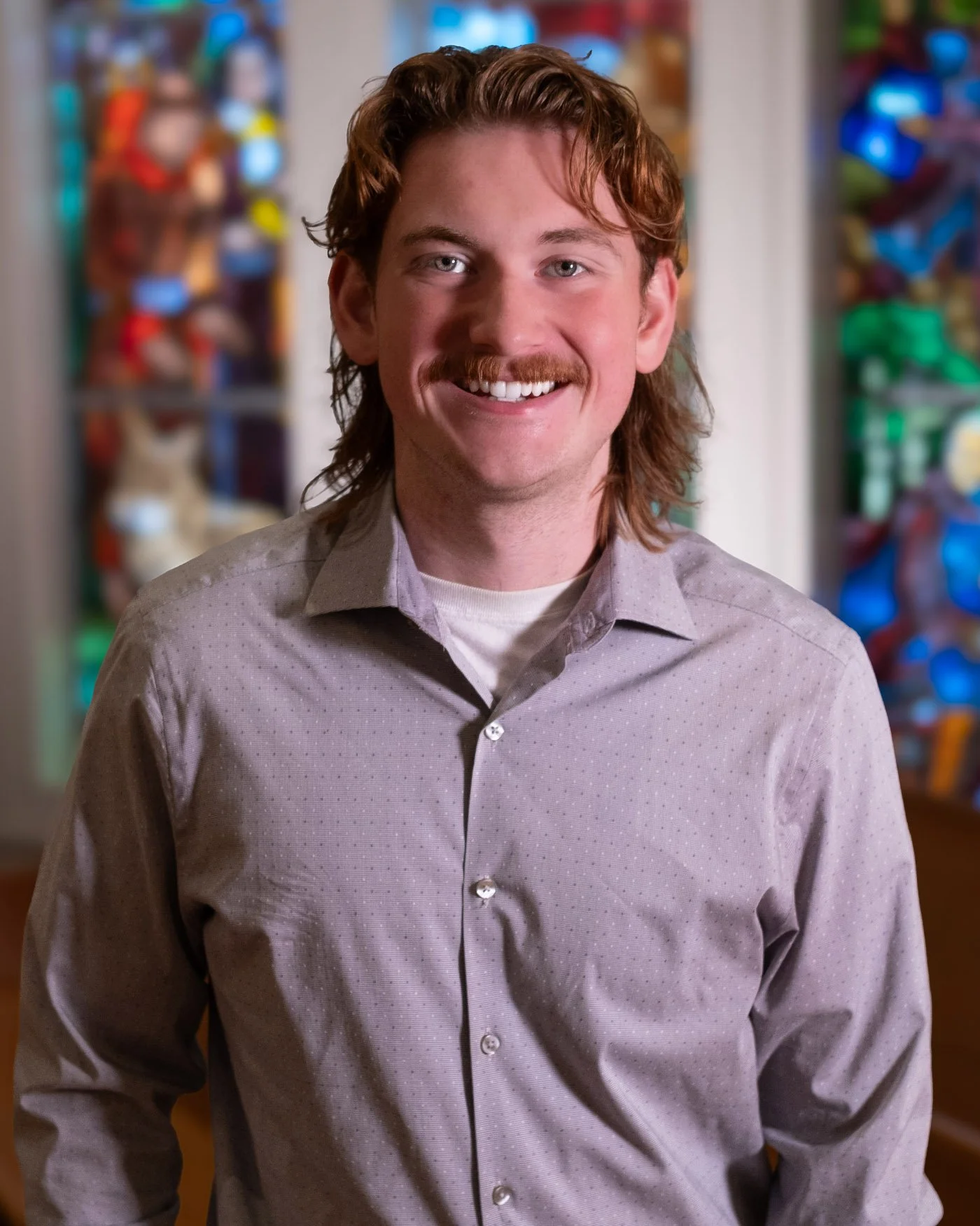 A man with red hair, a mustache, and a light purple shirt smiling in front of a colorful background.