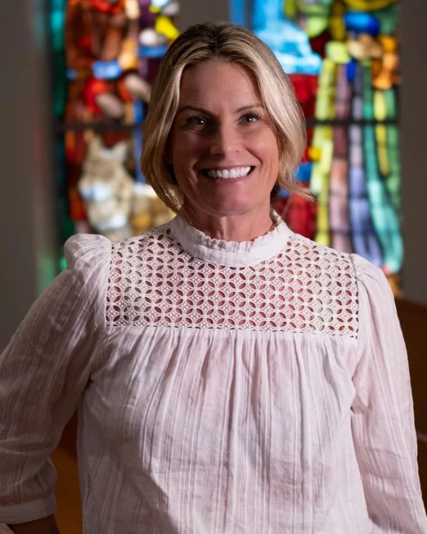 A smiling woman with shoulder-length blonde hair wearing a cream-colored sweater with navy horizontal stripes and decorative buttons on the shoulder, standing indoors with a background of windows and blurred interior elements.