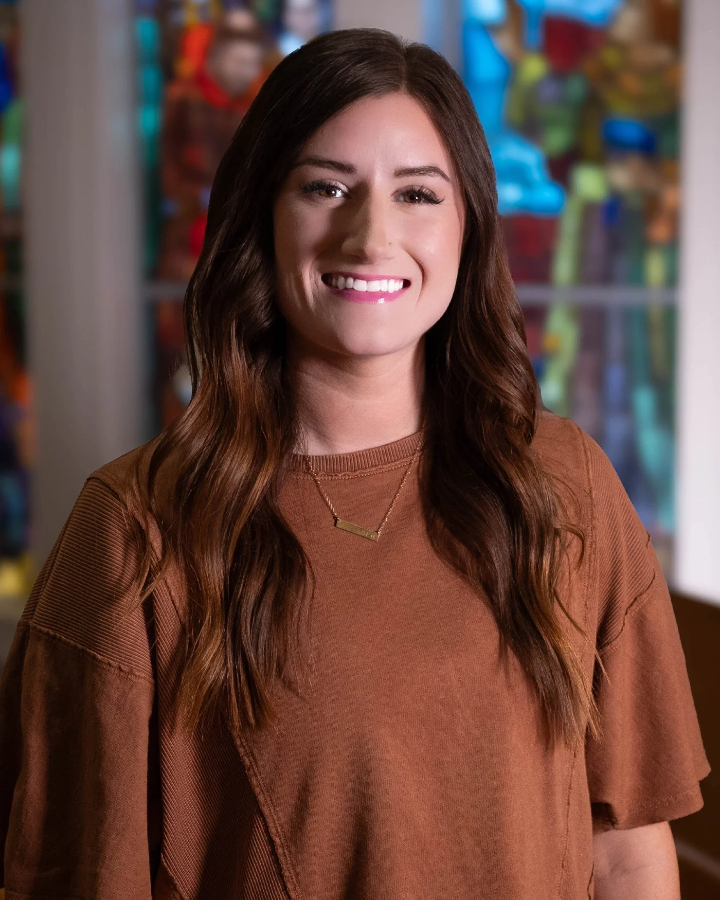 A woman with long wavy brown hair smiling, wearing a brown shirt and a gold necklace, standing indoors with blurred colorful objects in the background.