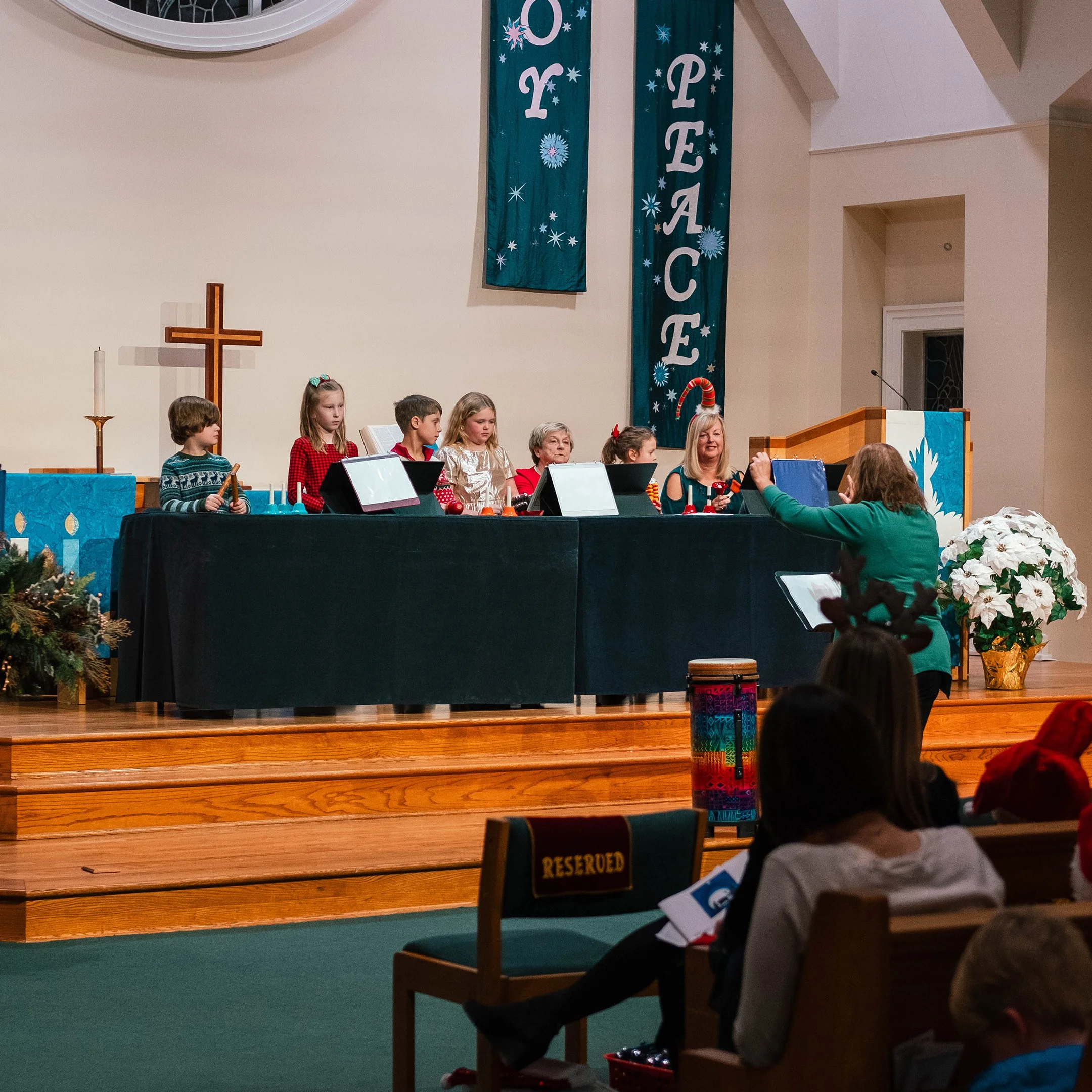 Children wearing Christmas attire perform on stage during a church Christmas program as their conductor leads them.