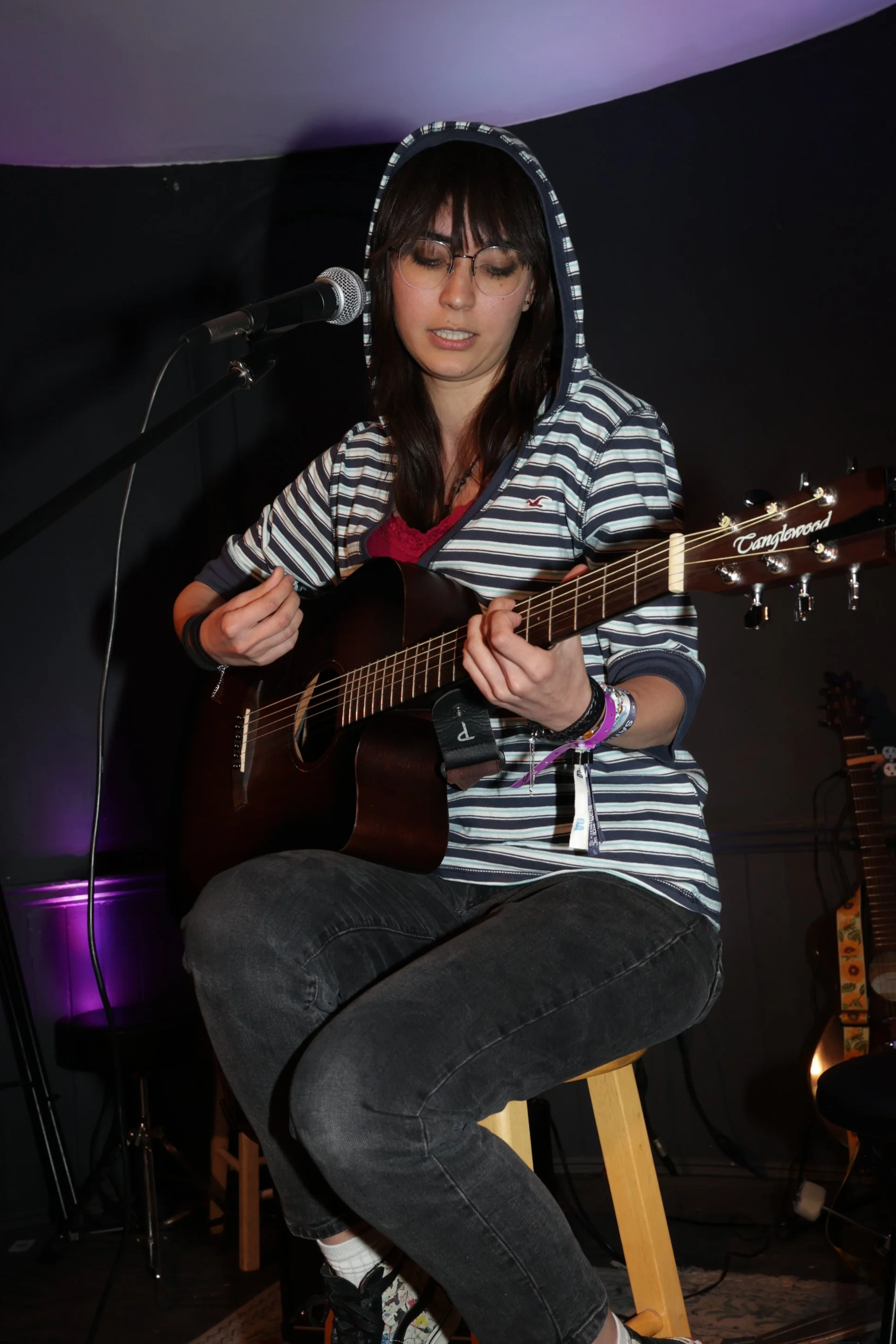 Kiara, a woman in a striped black and white hoodie, plays the guitar on stage.