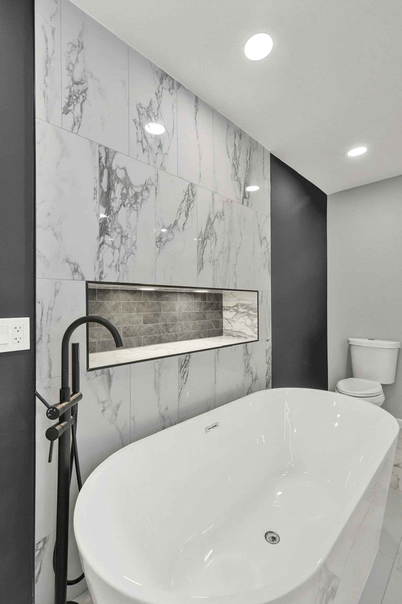 Modern bathroom with a white freestanding bathtub, marble tiled wall, and a toilet in the background.