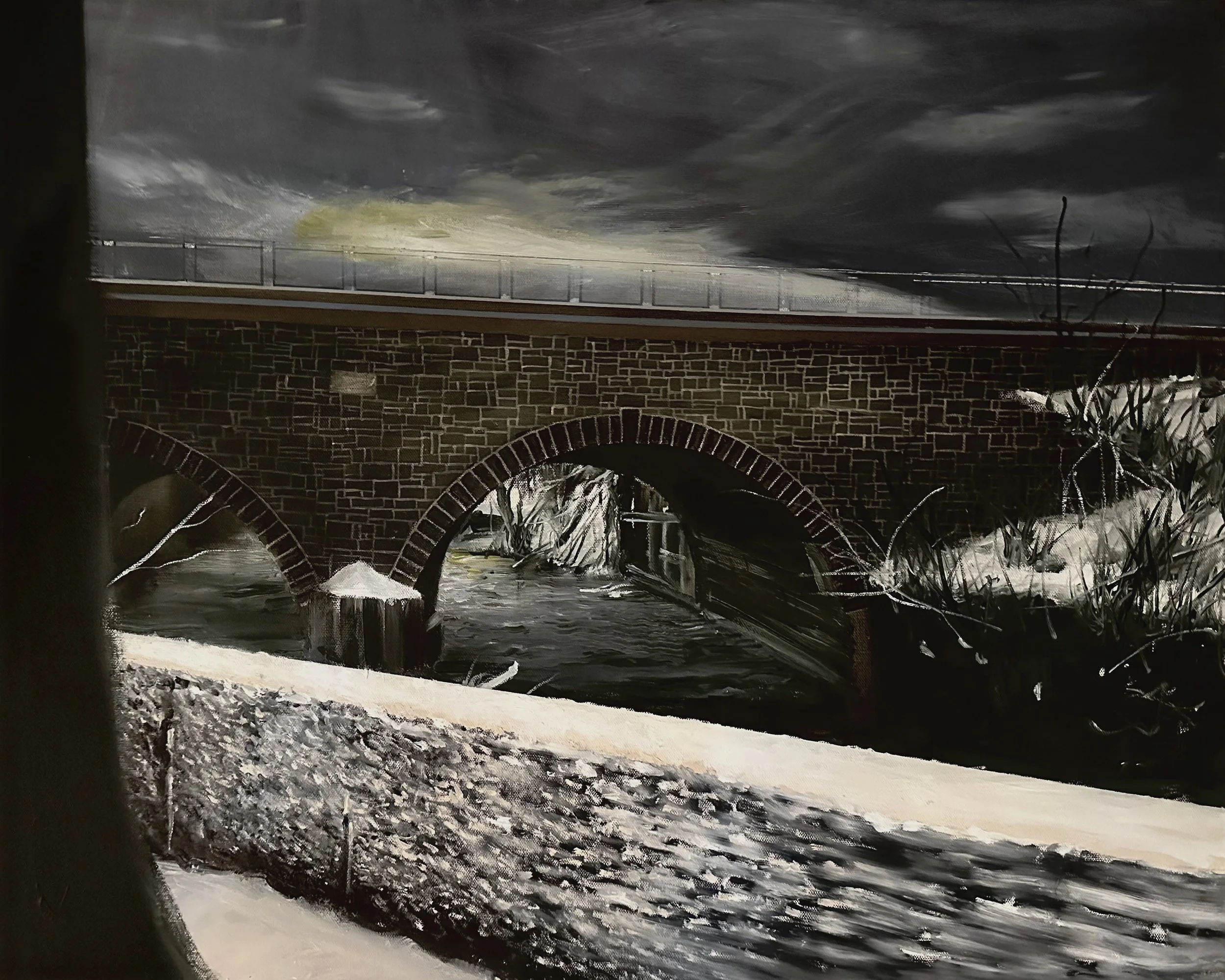 Nighttime scene of a brick bridge with an arched opening over a textured, snow-covered ground, dark cloudy sky, leafless trees, and fencing on top of the bridge.