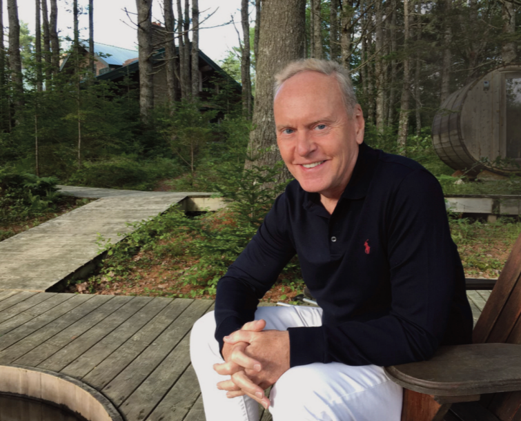 A smiling man with gray hair, wearing a navy blue long sleeve shirt and white pants, sitting on a wooden outdoor chair in a wooded area.