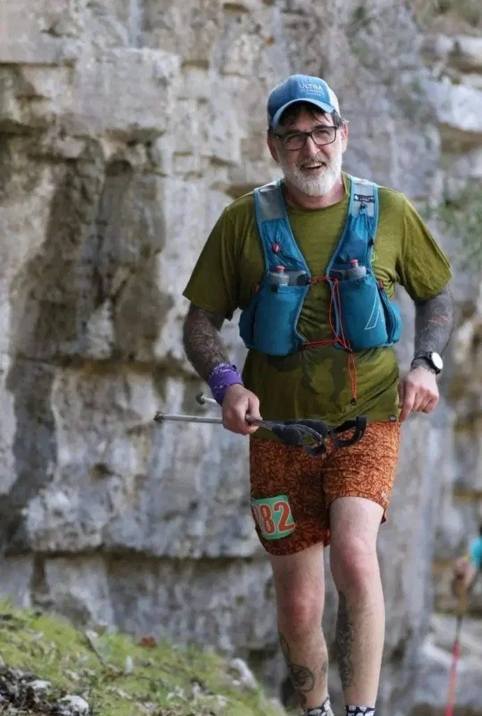 A smiling man in a green shirt, orange shorts, blue vest, and cap runs on a rocky trail, holding trekking poles. The setting is rugged and outdoors.