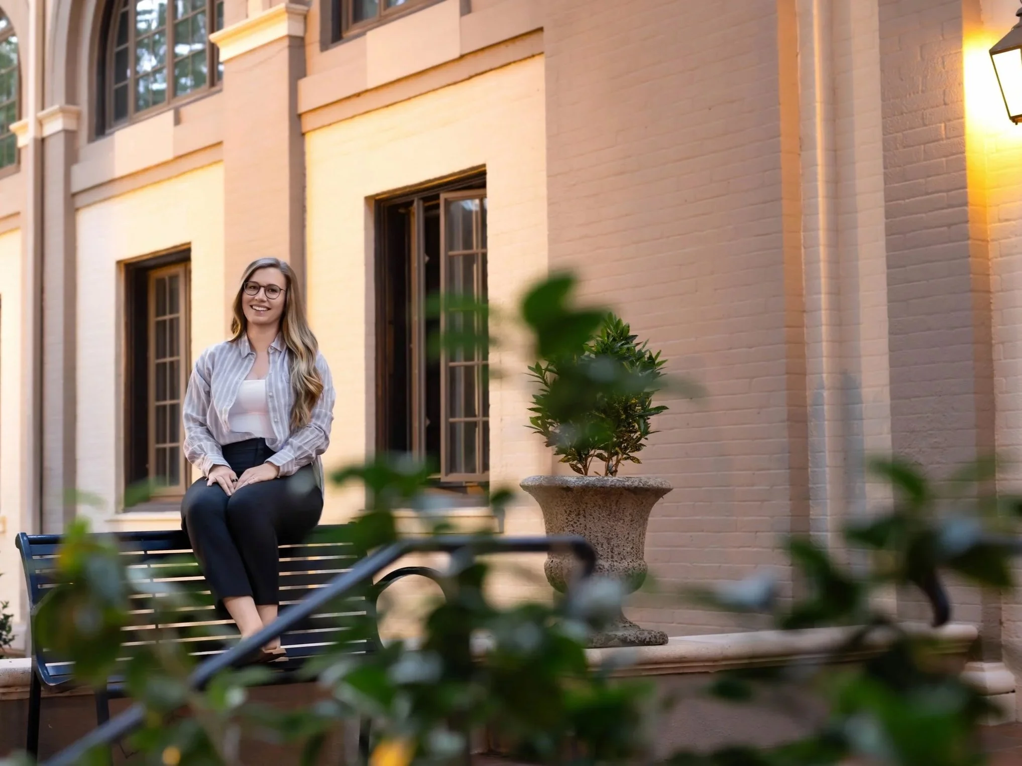 A smiling woman with long blonde hair, glasses, wearing a light-colored striped shirt and black pants, sitting on a park bench in front of a beige brick building, with a potted plant and a blurred foreground of green leaves.