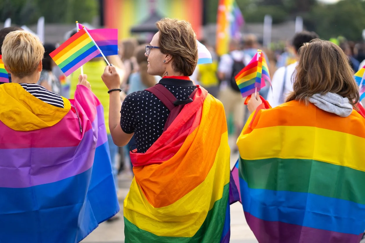 People participating in an LGBTQ pride parade holding rainbow flags, with some people wrapped in rainbow-themed flags.