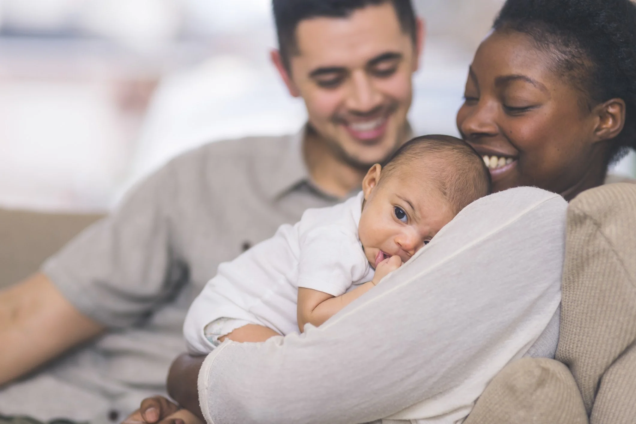 A diverse family holding a baby, with the baby lying on the mother's shoulder while she smiles and father looks on, in a cozy home setting.
