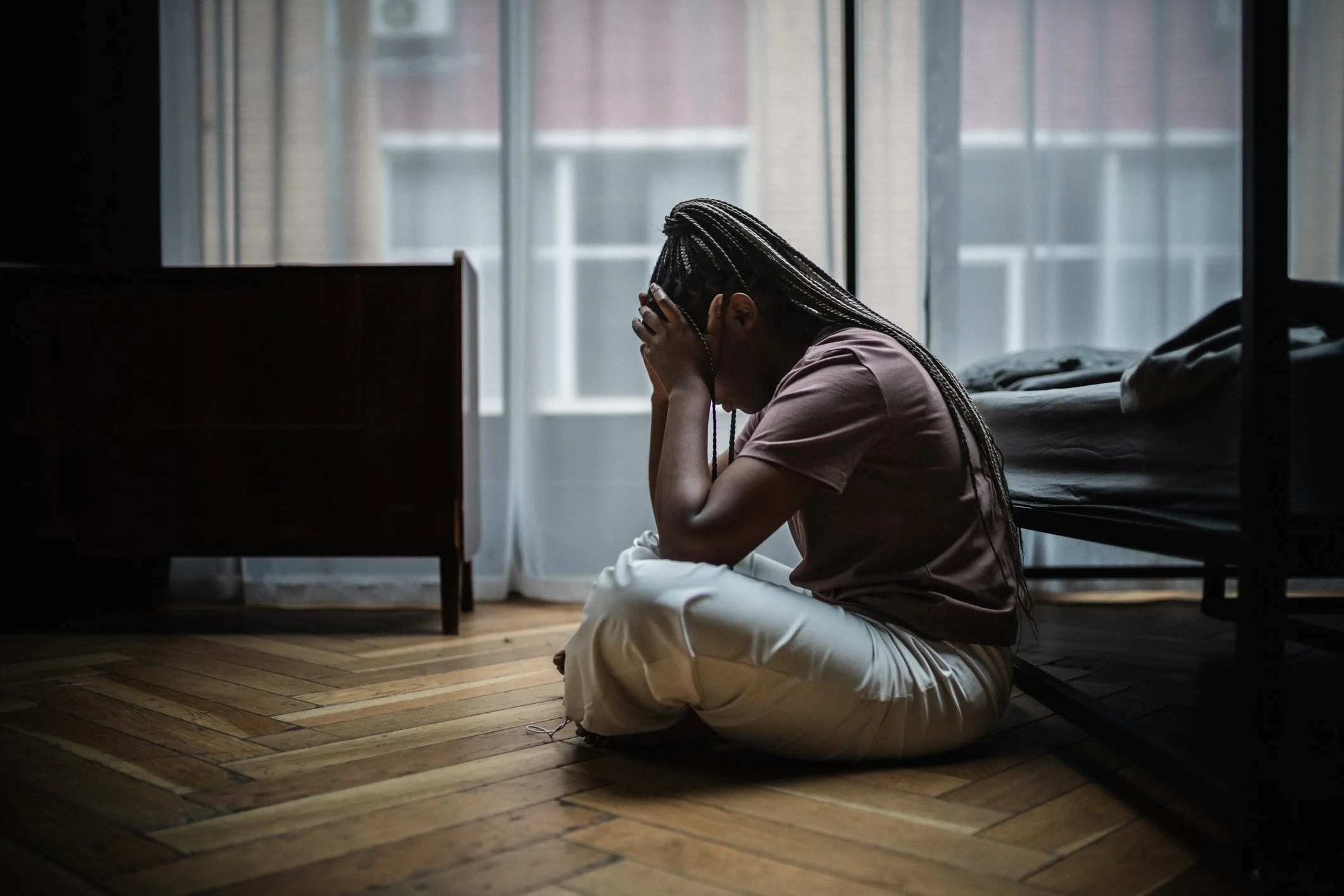 A woman sitting on the floor with her knees up, holding her head in her hands, in a dimly lit room with natural light coming through the window.