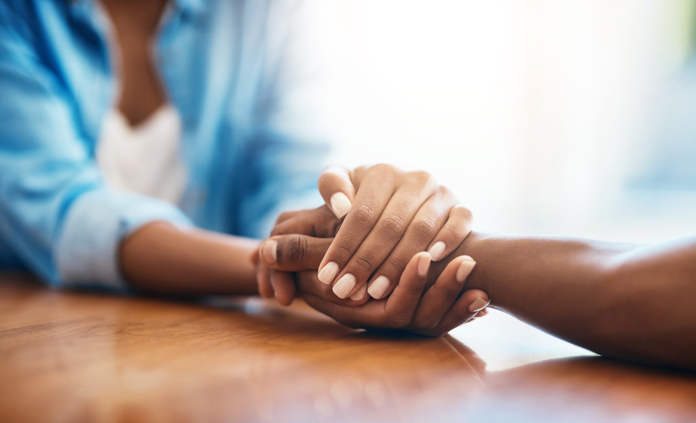 Two people holding hands on a wooden table, with one person's hand resting gently on top of the other's.