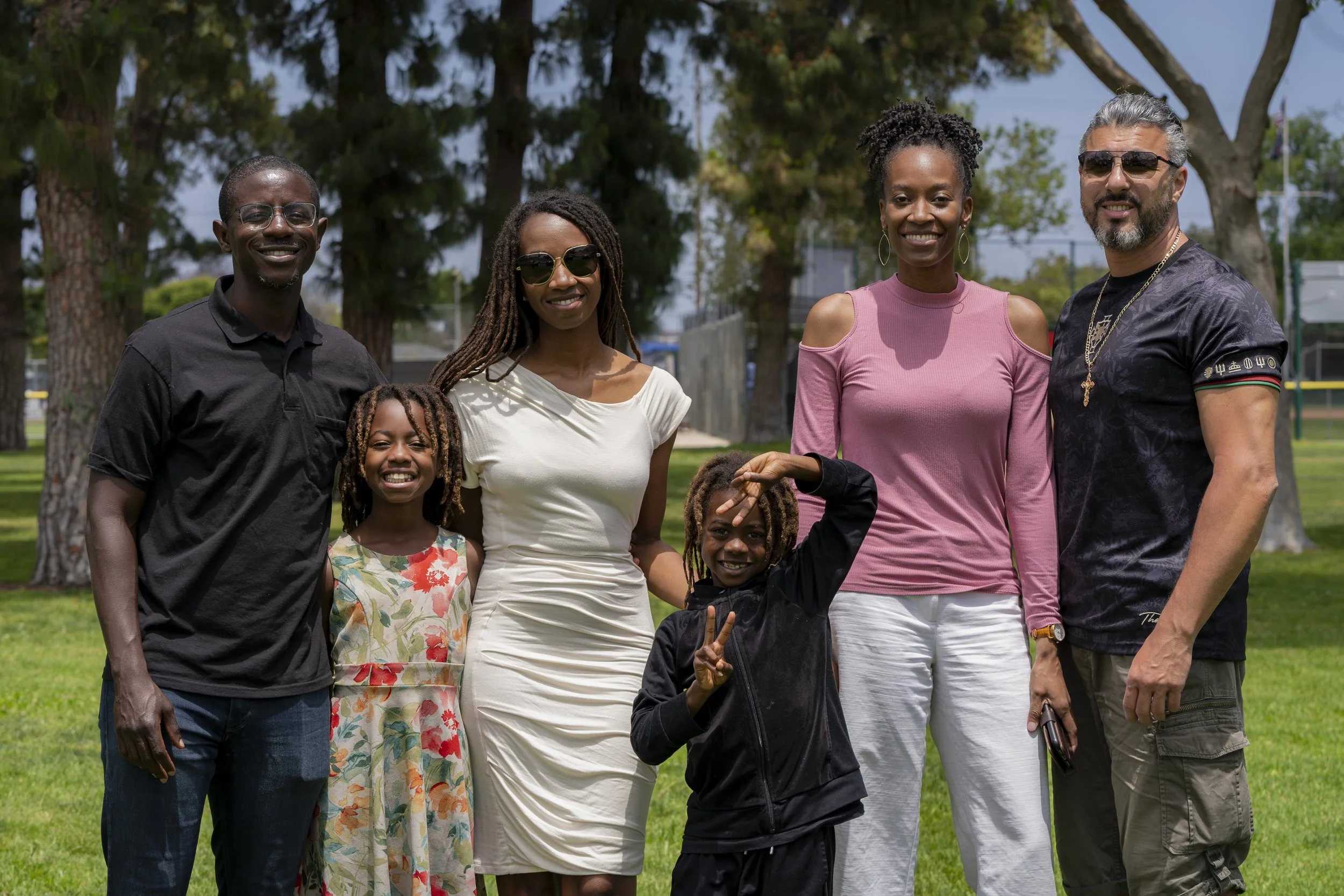 Group of six diverse people standing outdoors in a park on a sunny day, smiling and posing for the camera.