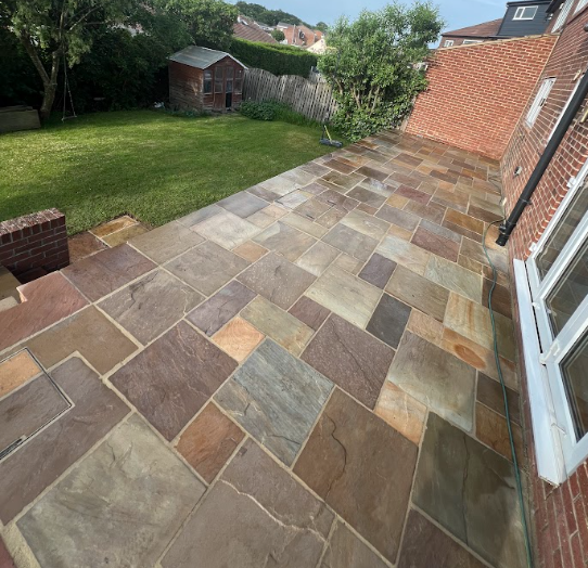 Backyard patio with multicolored stone pavers, bordered by a brick house wall, with a lawn, wooden shed, trees, and surrounding houses visible in the background.