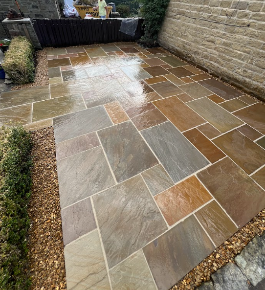 Wet multicolored stone patio in a backyard, bordered by small bushes and a stone wall.