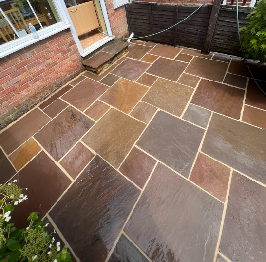 Newly installed stone patio with brown, beige, and gray rectangular and square paving stones outside a house, with a brick wall, window, and green plants nearby.