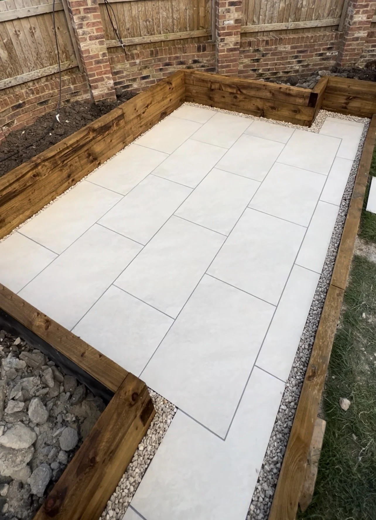 A backyard patio with light-colored tiles, bordered by wooden planks and small white gravel, with a brick wall and wooden fence in the background.
