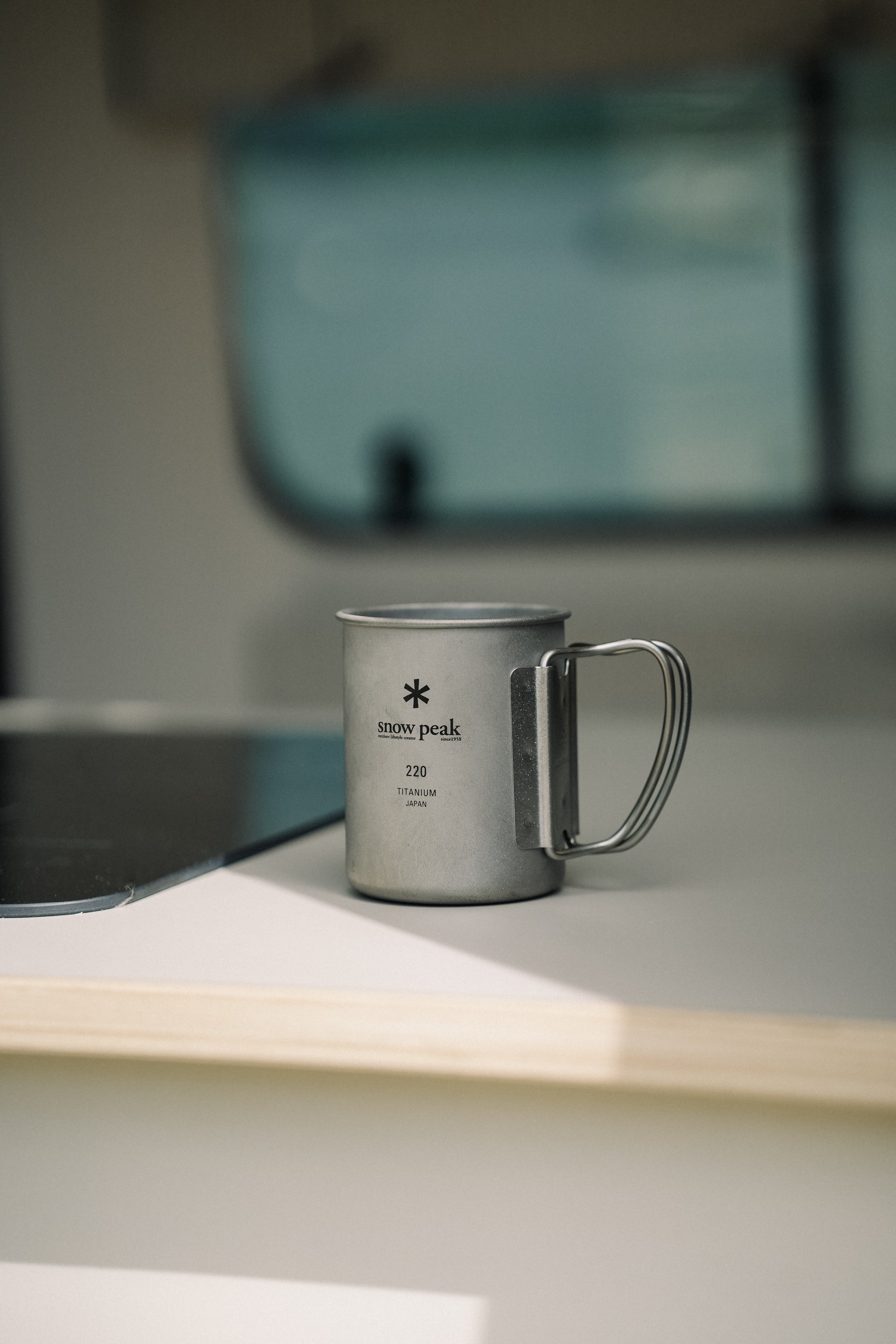 A small titanium Snow Peak mug with a handle on a white surface, with a blurred background.