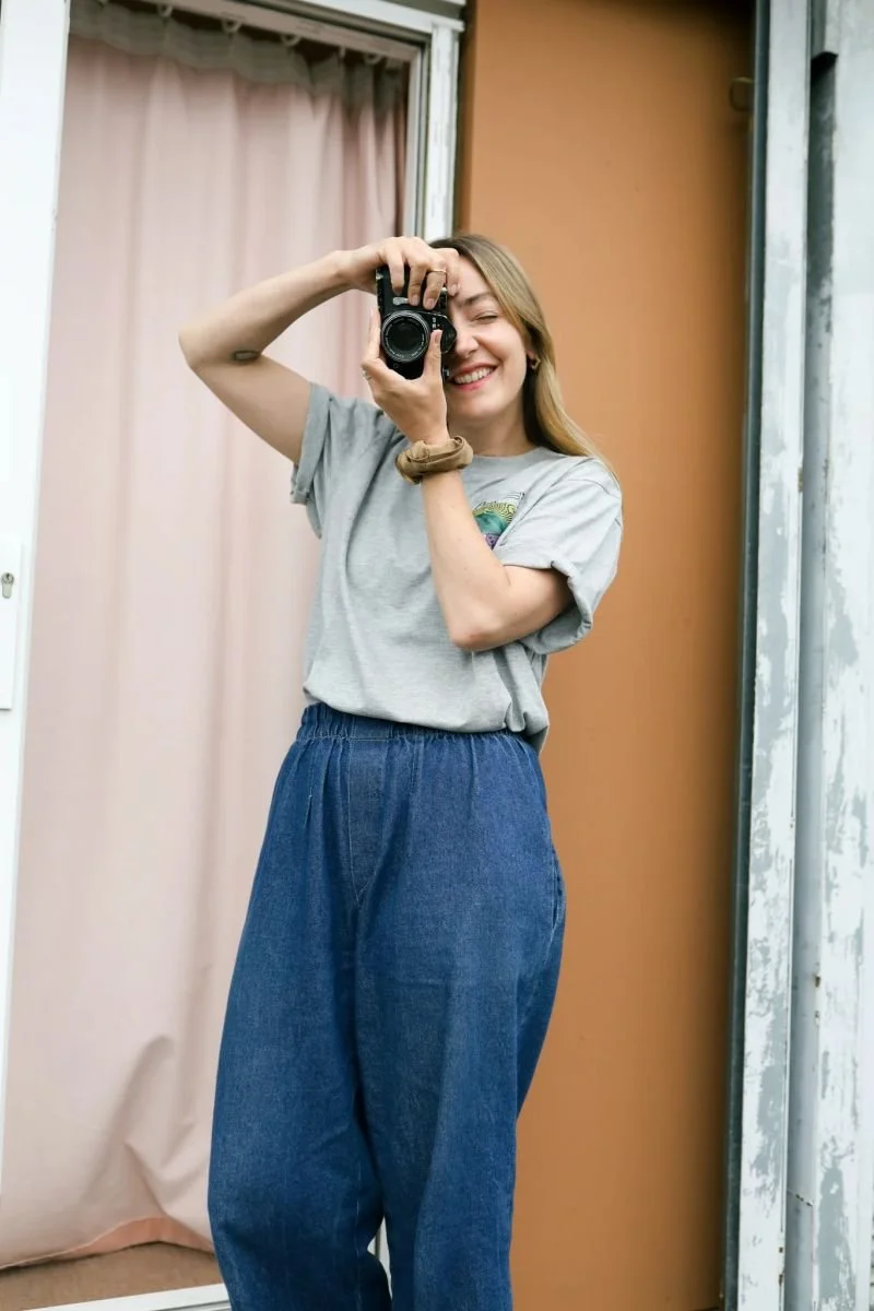 A young woman smiling and taking a selfie with a camera in front of a mirror. She is wearing a gray T-shirt and loose blue jeans, with her hair down.