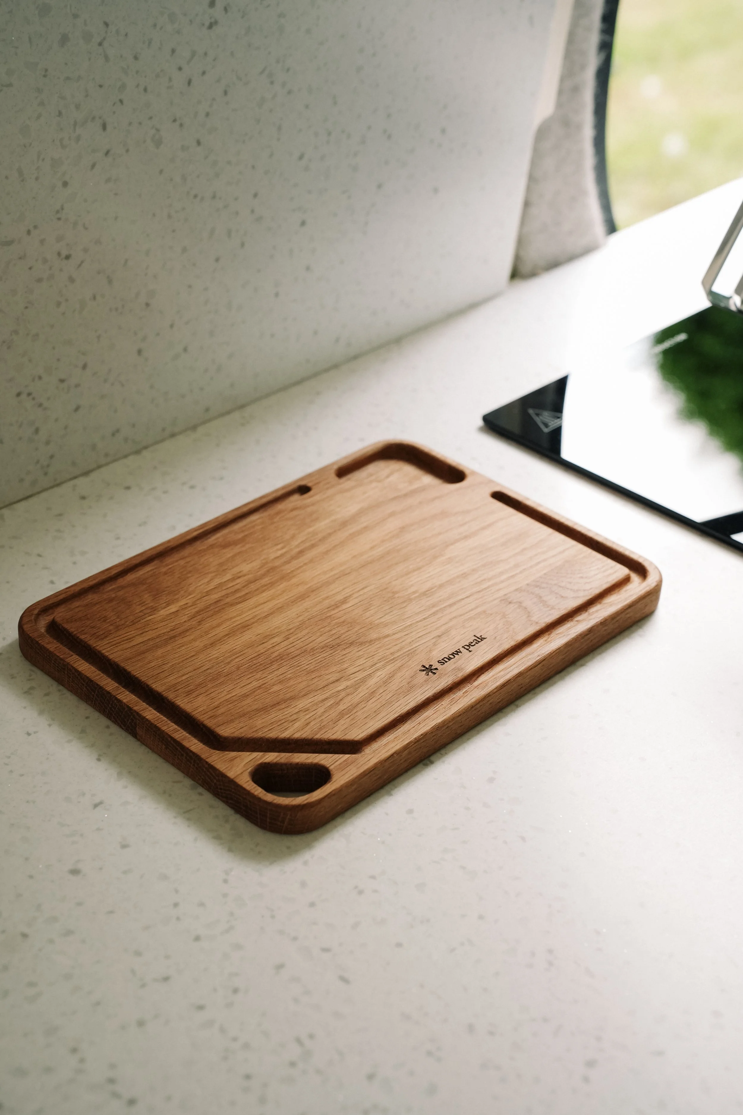 Wooden cutting board on a white speckled countertop with a reflective stove top and a window in the background.