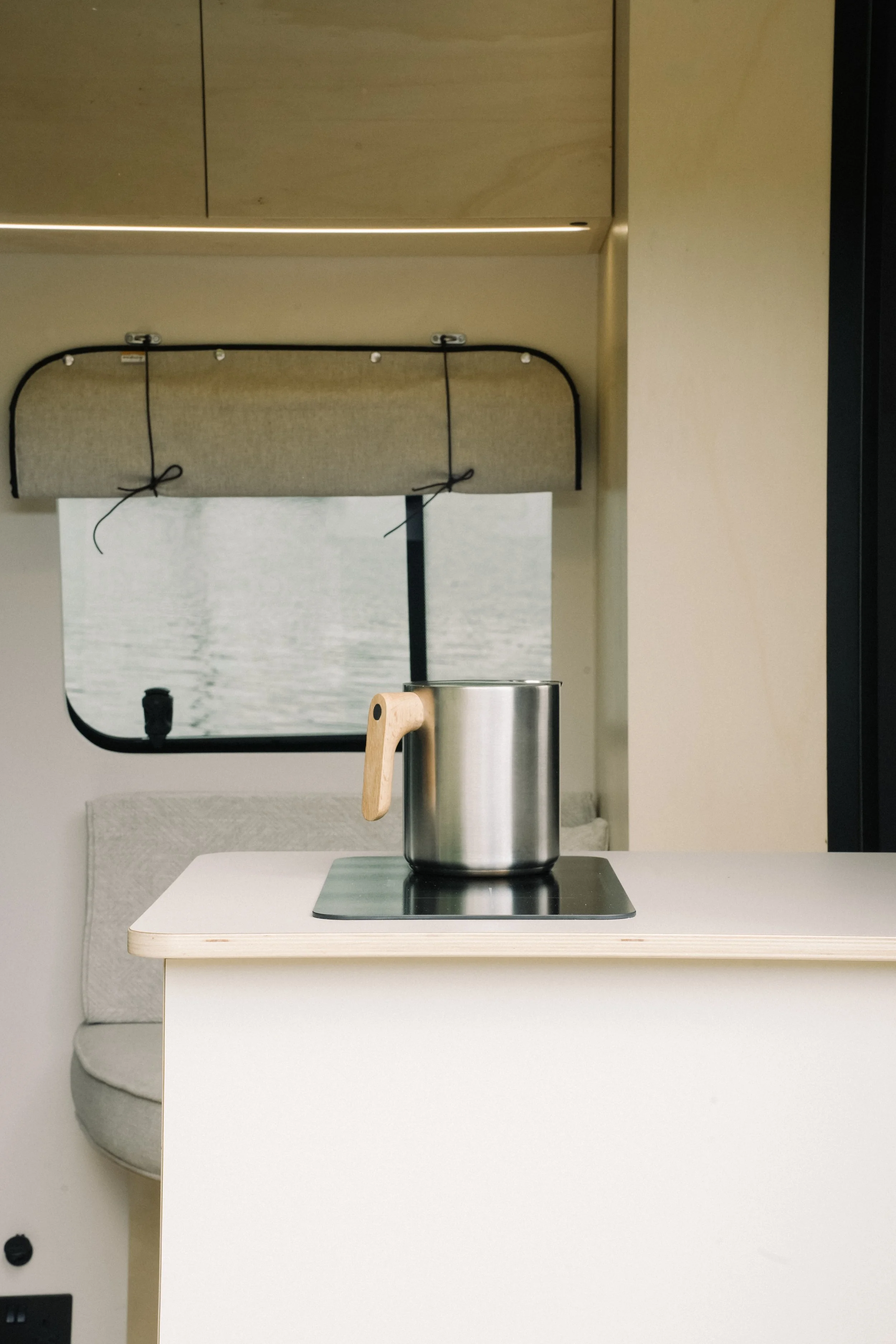 A metal cooking pot with a wooden handle on a stovetop inside a minimalist trailer or camper kitchen, with a window showing water outside.