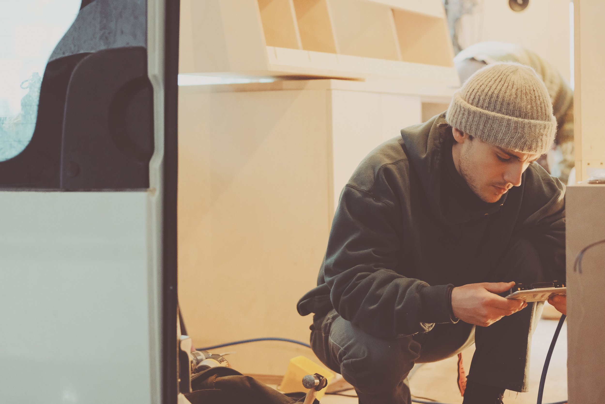 A young man in a gray knit cap and black hoodie squats while working on an electronic device or project, surrounded by tools and materials in a workshop or carpentry space.