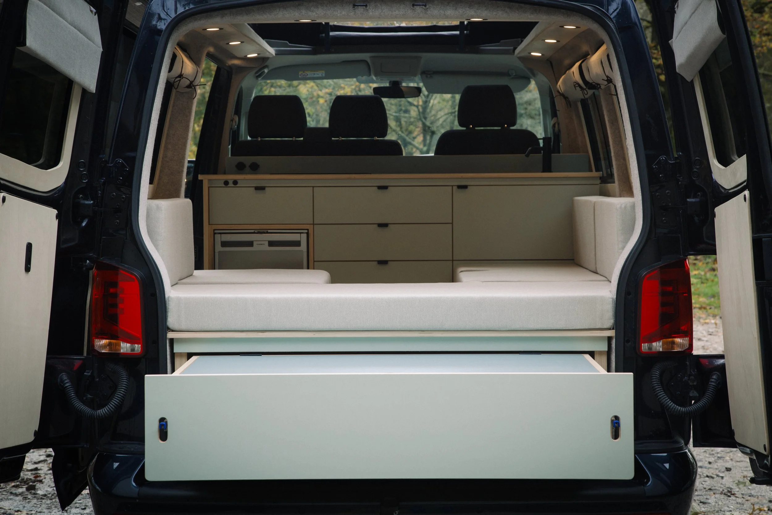 The back of a black van with open rear doors showing a built-in camper with beige seating, storage drawers, and a small refrigerator, set outdoors in a natural setting.