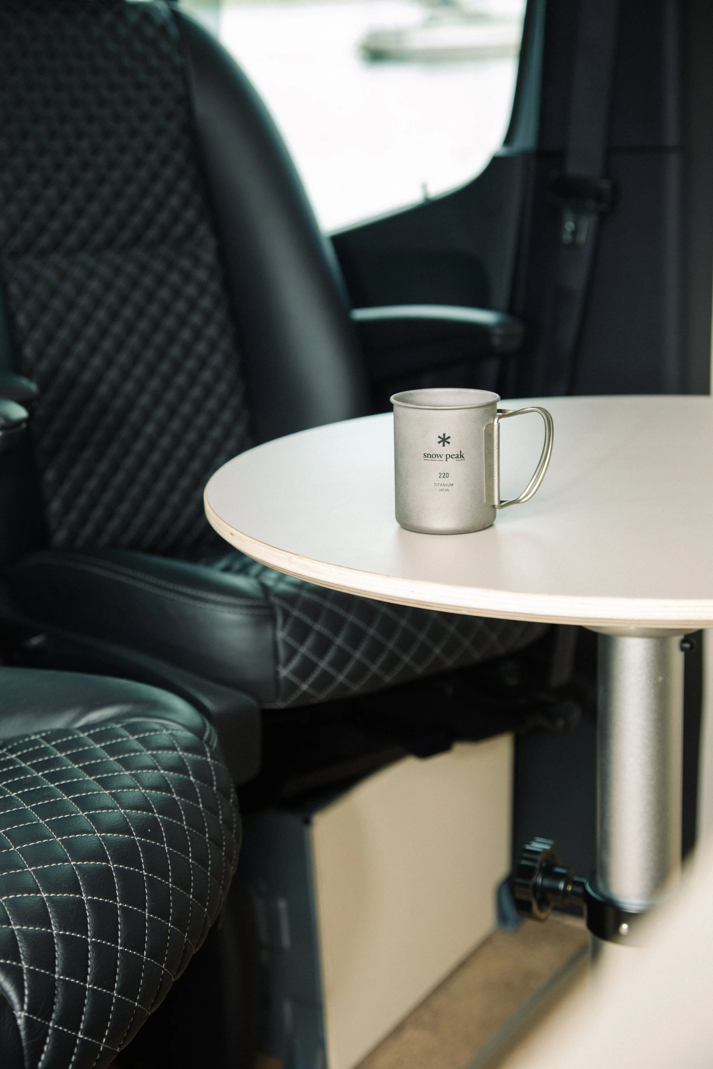 A coffee mug on a tiny round white table inside a vehicle, with black quilted seat upholstery visible, suggesting a cozy and modern interior.