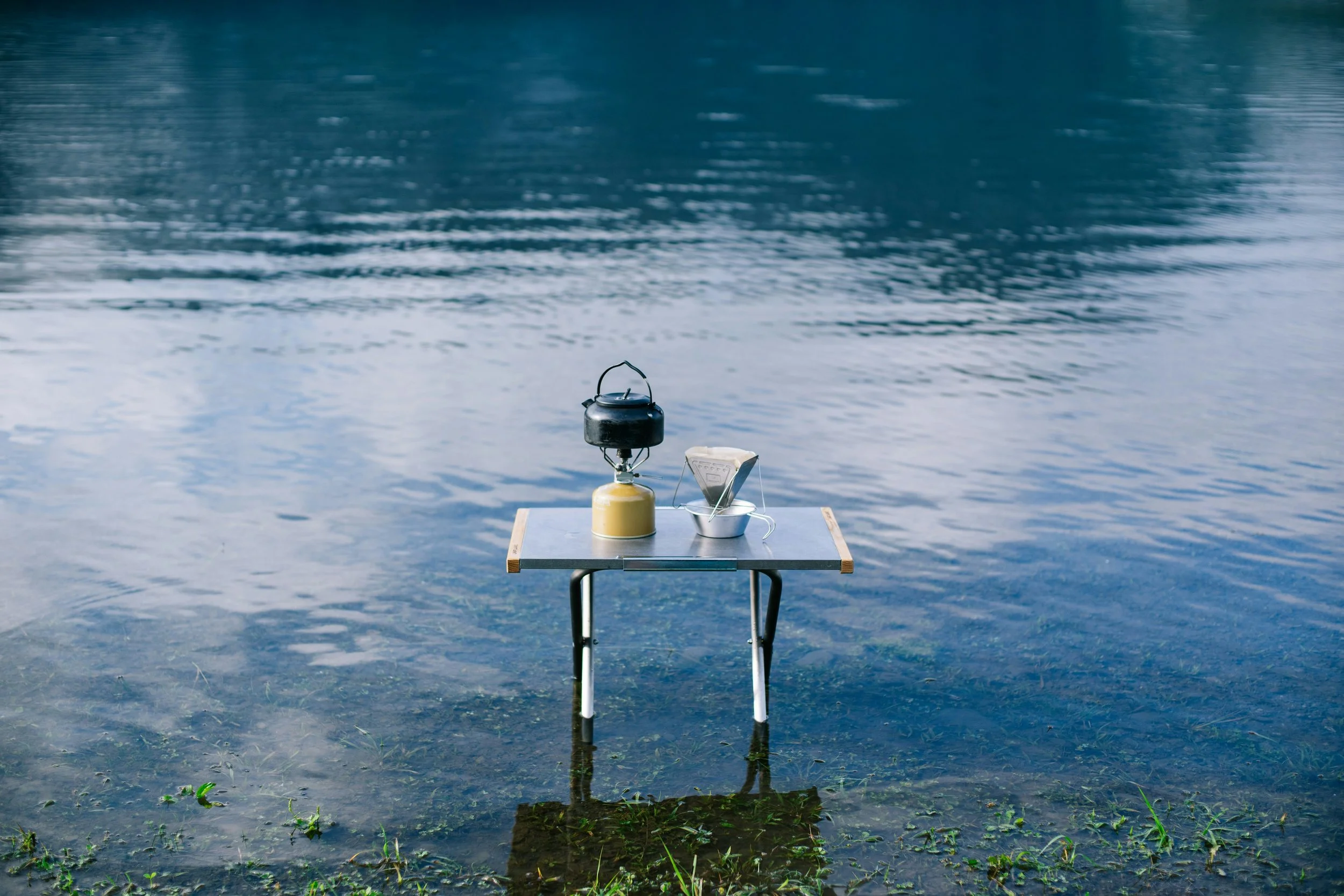 A small table with a camping stove, a bowl of face masks, and a water bottle, set up in shallow water near a lake.