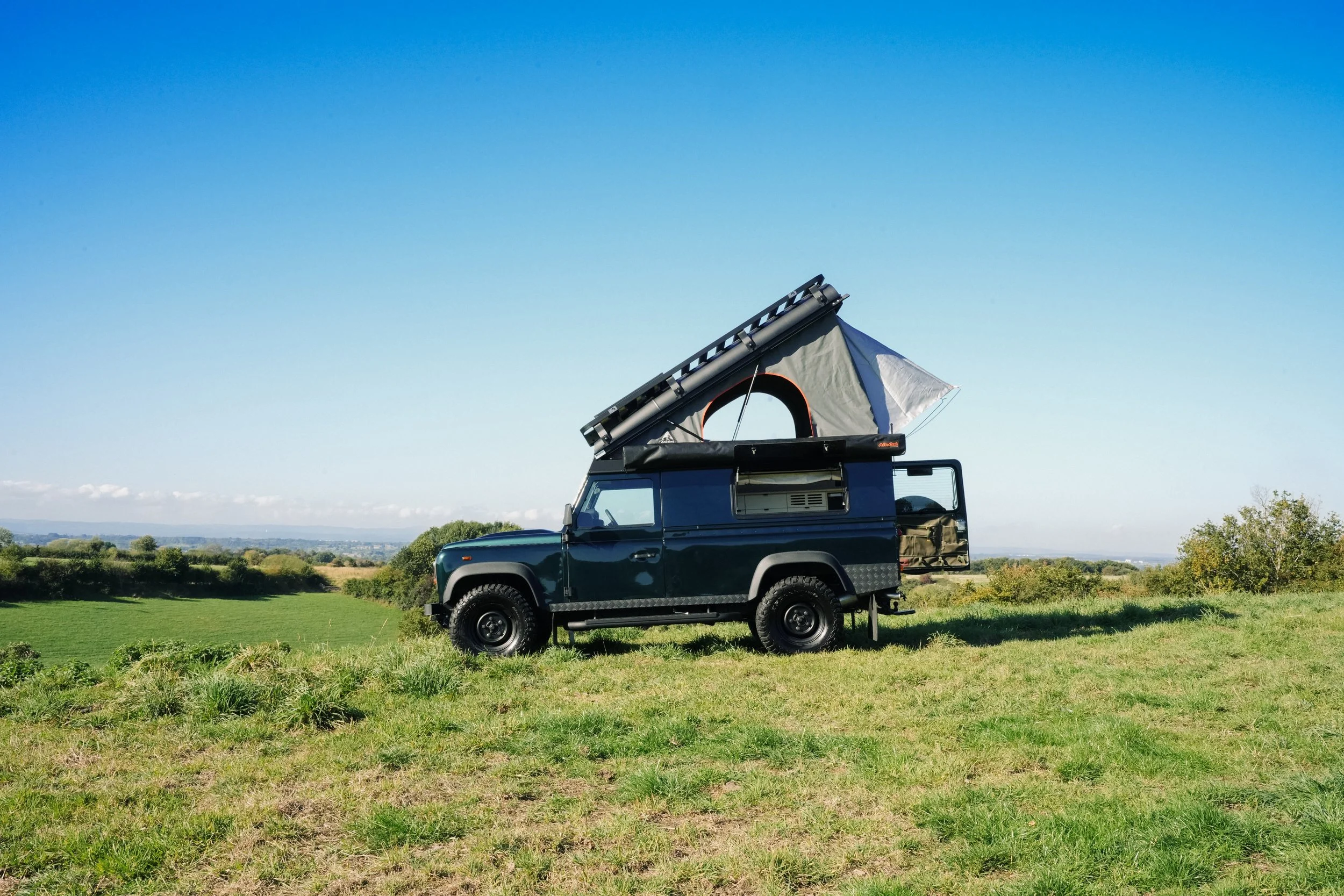 A black off-road vehicle with a rooftop tent and solar panel set up on a grassy field under a clear blue sky.