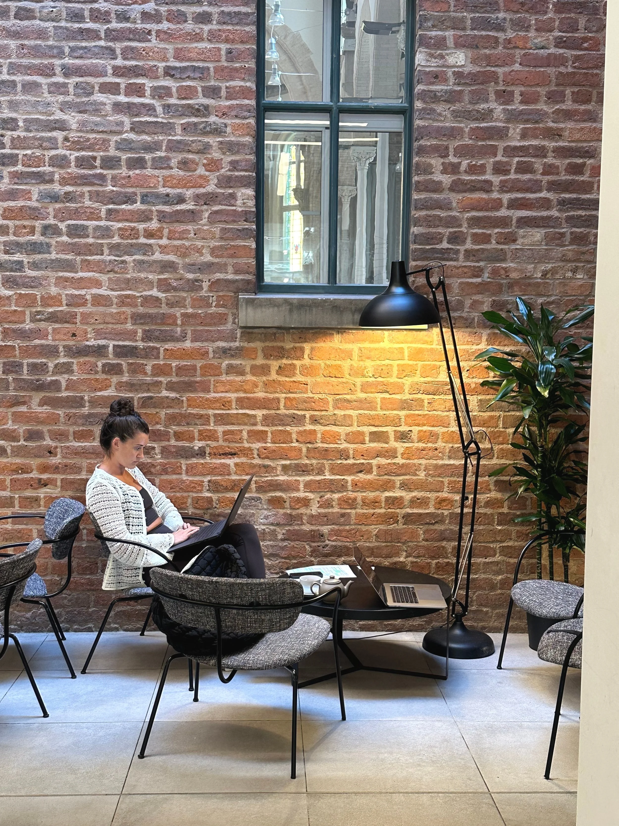 A woman sitting at a table with a laptop, coffee cups, and a phone, in a room with an exposed brick wall, tall window, and a floor lamp.
