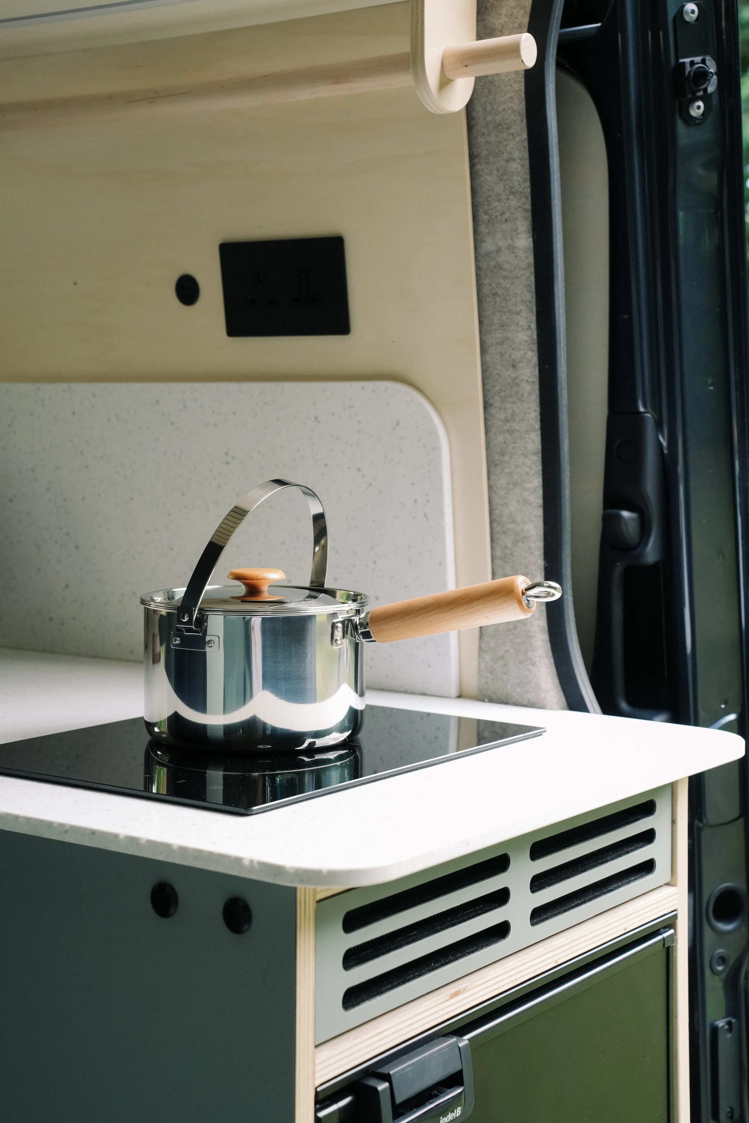 A small kitchen setup inside a vehicle with a black electric stove, a shiny stainless steel pot with a wooden handle, a white speckled countertop, a power outlet, and part of a window.