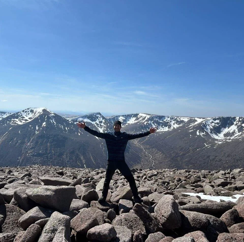 A person standing on rocky terrain with snowy mountain peaks in the background under a blue sky.