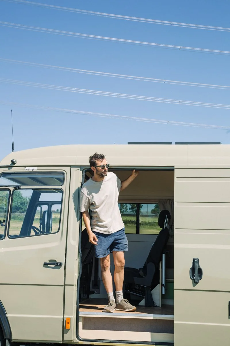 A man standing inside a camper van, leaning out of the side door, with a scenic green field and blue sky with power lines in the background.