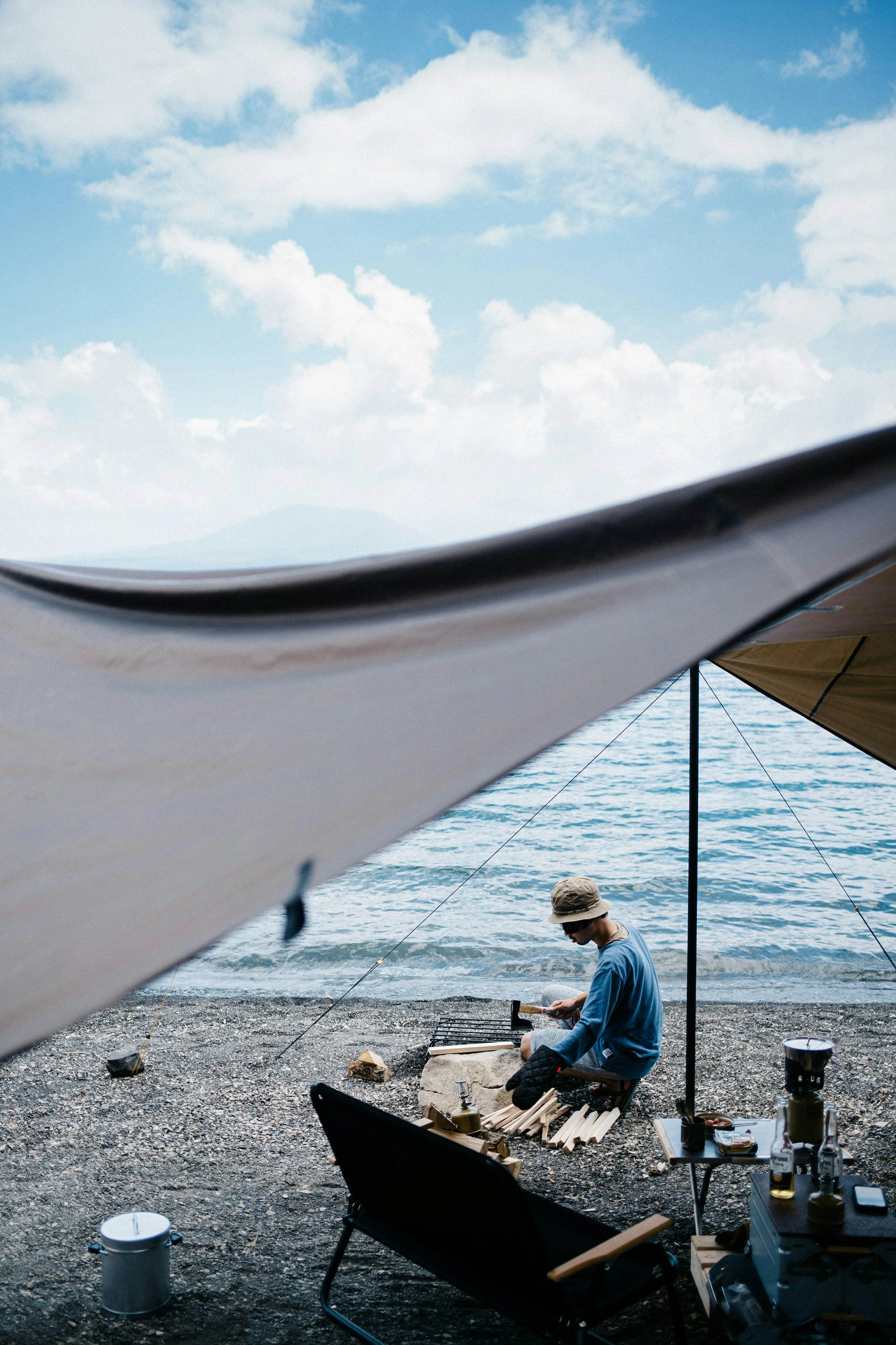 Person sitting on a small stool by the rocky beach, grilling food and chopping wood under a large tent near the water, with a sailboat canopy overhead and a mountainous landscape in the distance under a partly cloudy sky.