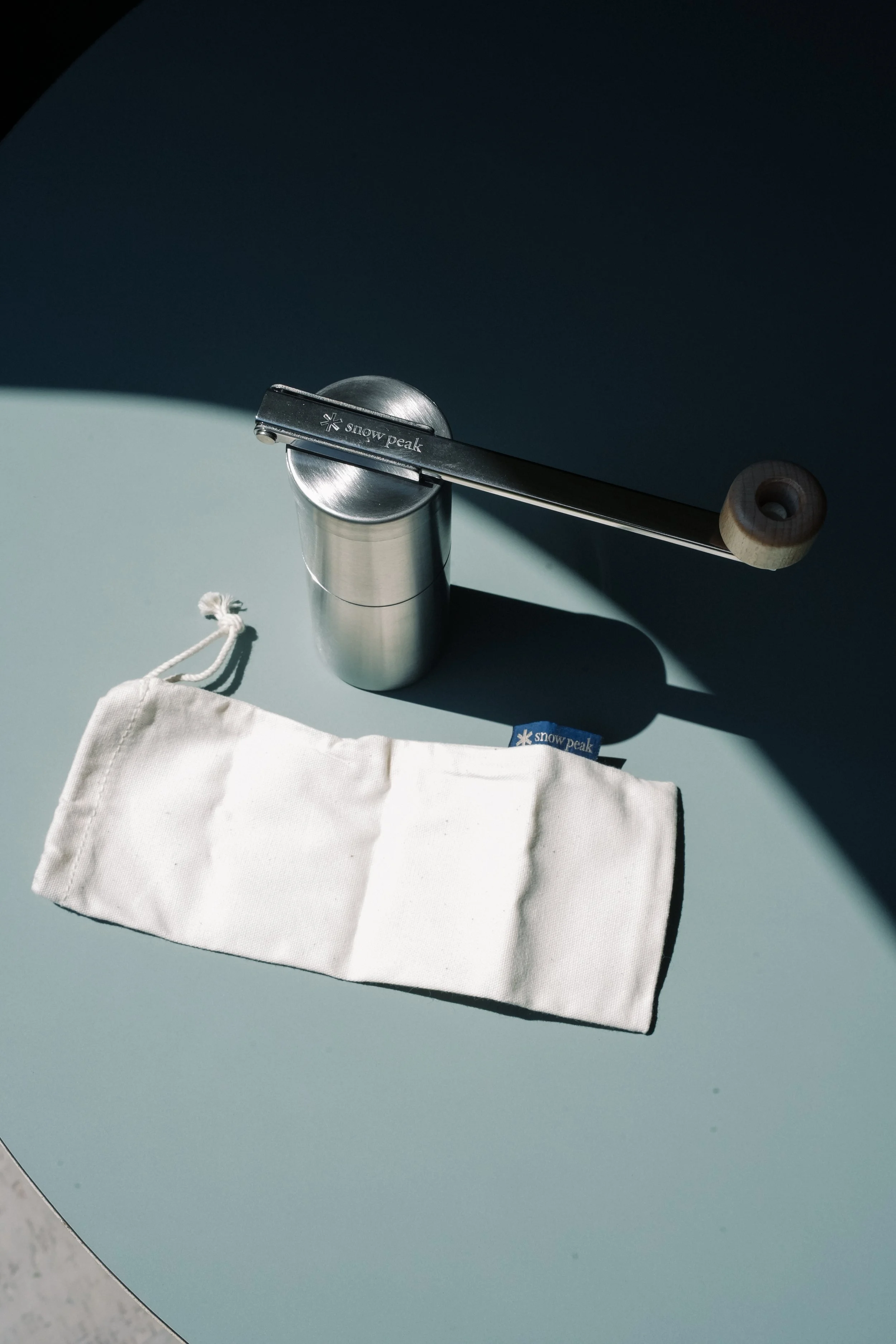 A silver pepper grinder, a wood-handled pepper mill, and a white fabric pouch with a 'snow peak' label on a light green surface, with shadows cast across the objects.