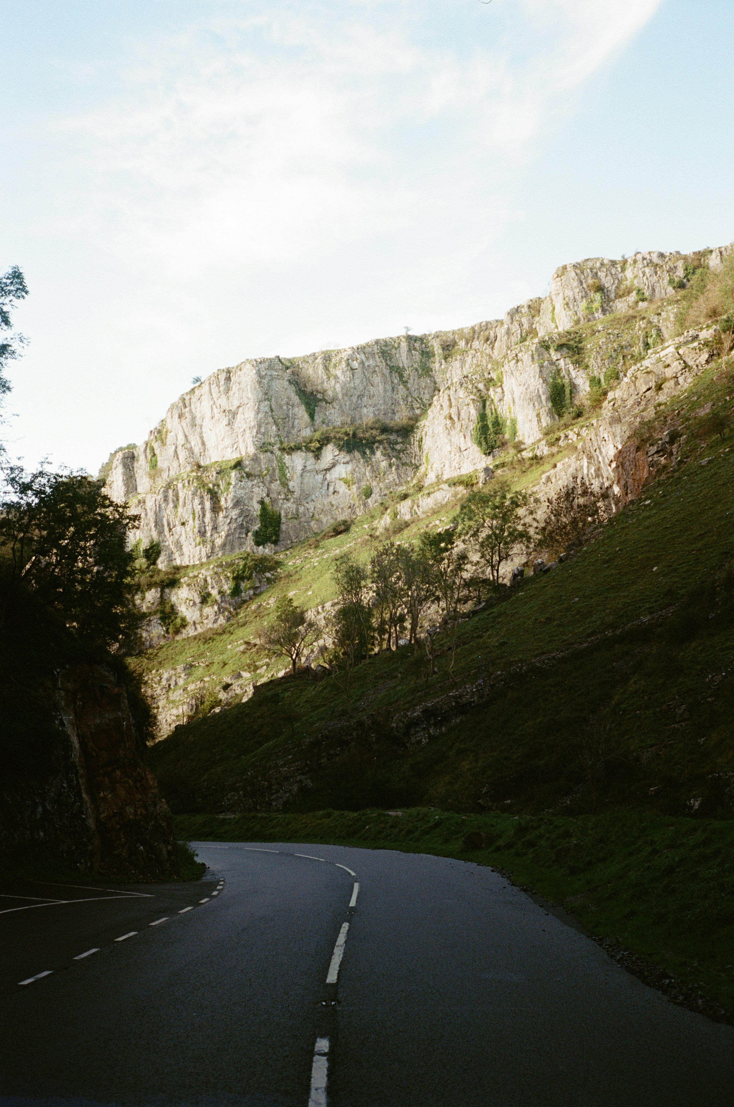 A winding mountain road with a steep rocky hillside and sparse trees, under a sky with scattered clouds.
