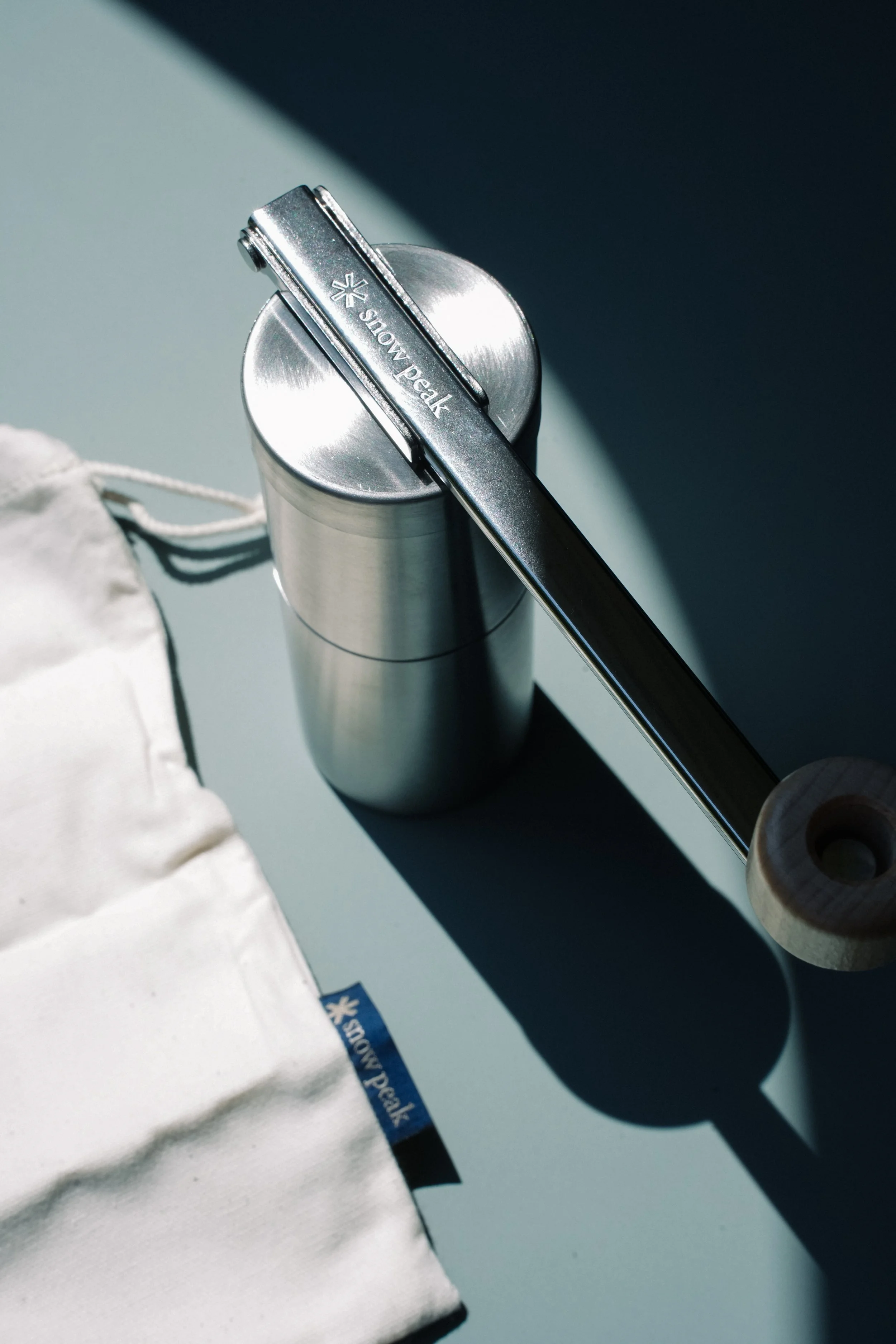 A metallic Snow Peak thermos with a logo, a wooden-handled utensil resting on top, and a white cloth item with a small blue Snow Peak tag, all on a light-colored surface with a shadow cast by sunlight.
