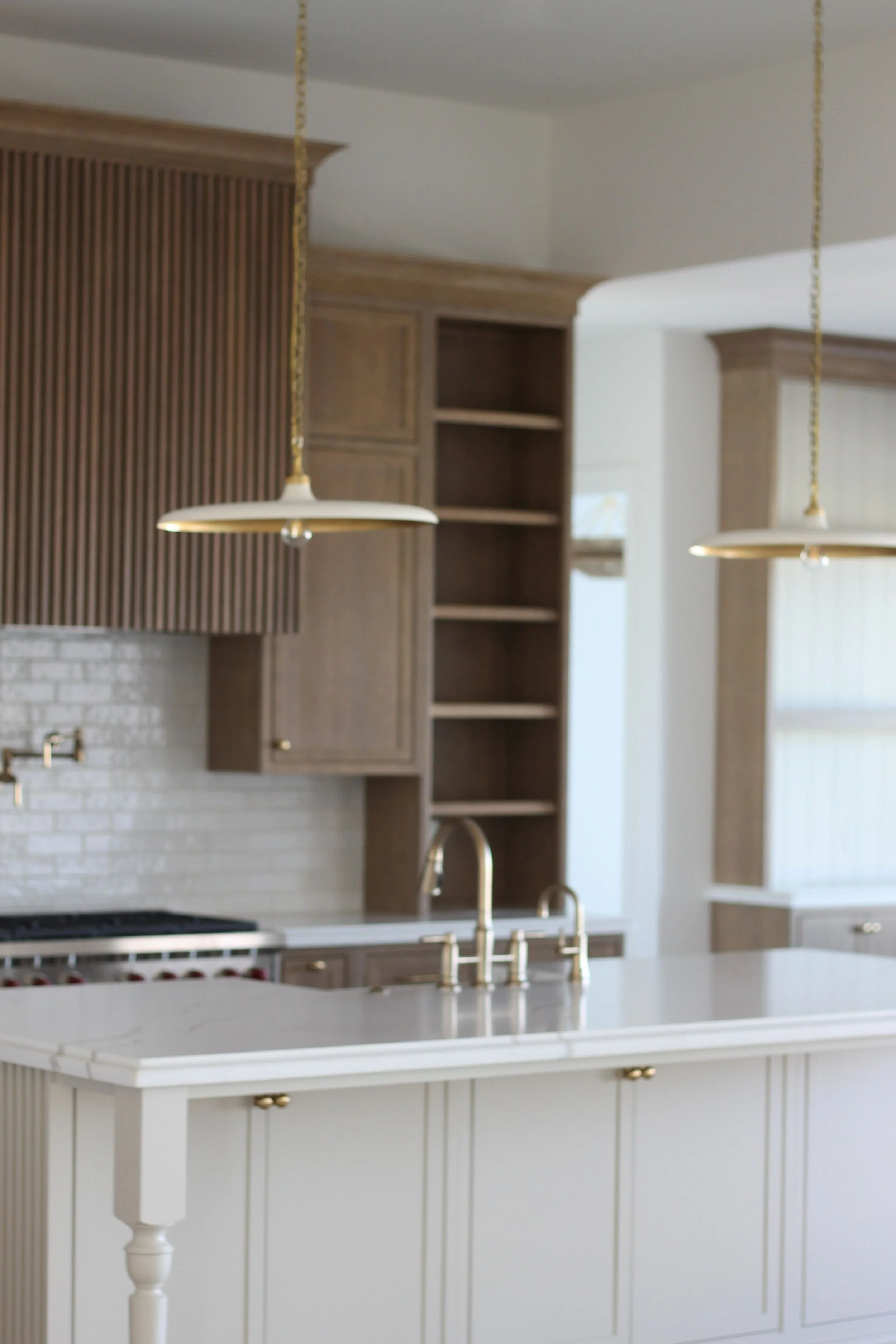 Modern kitchen with white island, wooden cabinetry, open shelves, gold hardware, and gold pendant lights.