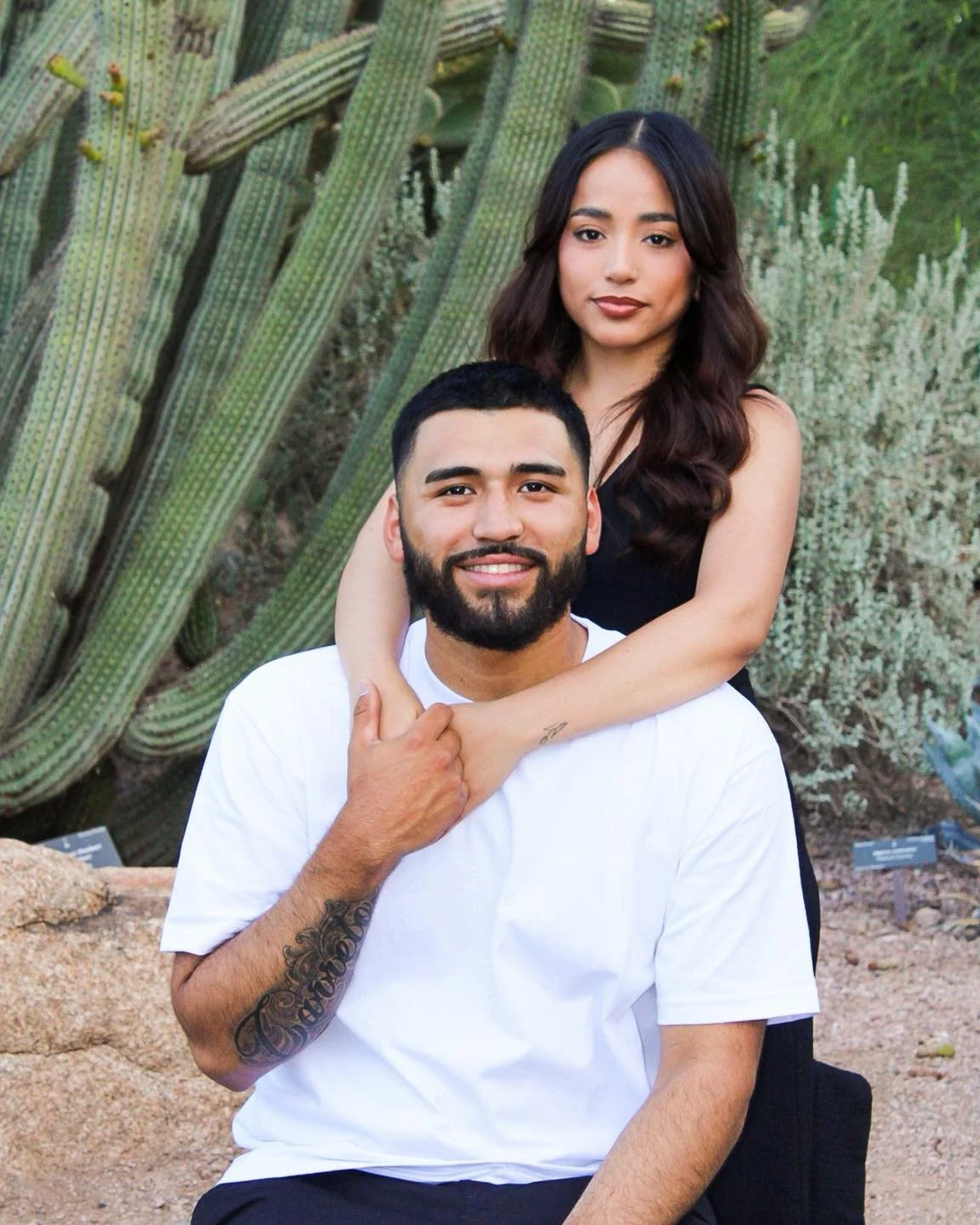 A young couple outdoors in front of large cacti, with the woman standing behind and resting her arms on the man's shoulders, both smiling at the camera.