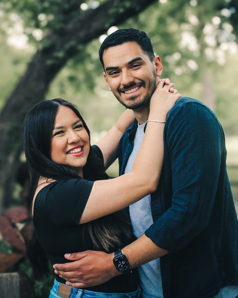 A happy couple embracing outdoors in a park with trees and green foliage in the background.