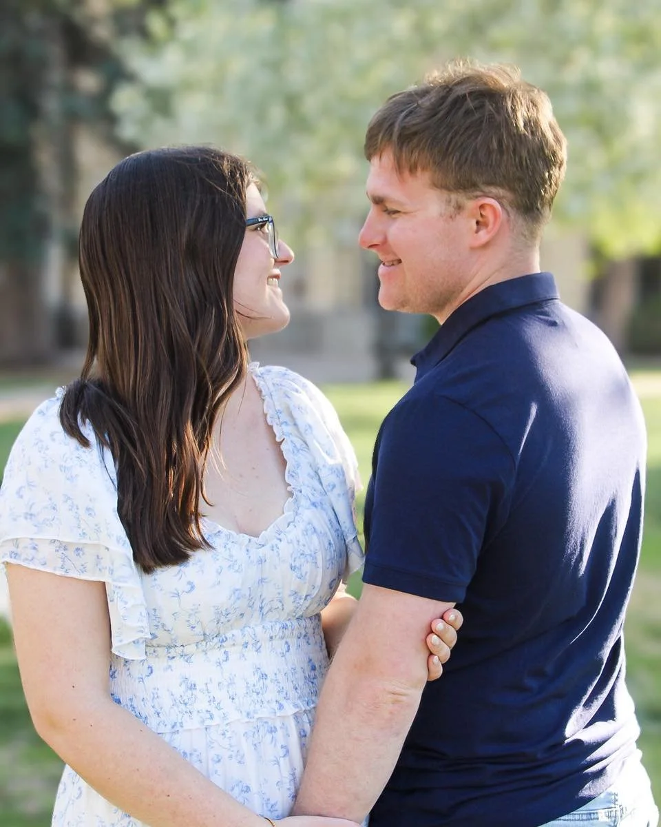 A couple standing close, smiling at each other outdoors on a sunny day.